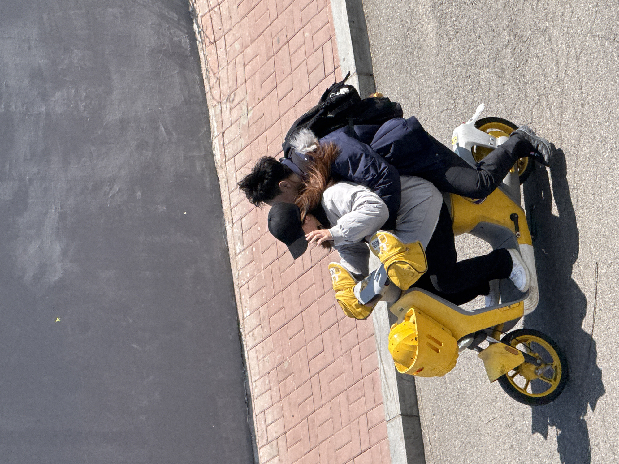 Aerial perspective of a yellow e-bike on a Zhongtun city street. Three individuals are piled on the e-bike near a sidewalk consisting of red bricks. One person wears a black cap and gray sweatshirt. The second individual wears black pants, a blue jacket, and black shoes. A third person is between them wearing yellow gloves. A black backpack is on the back of the e-bike. The street is asphalt.