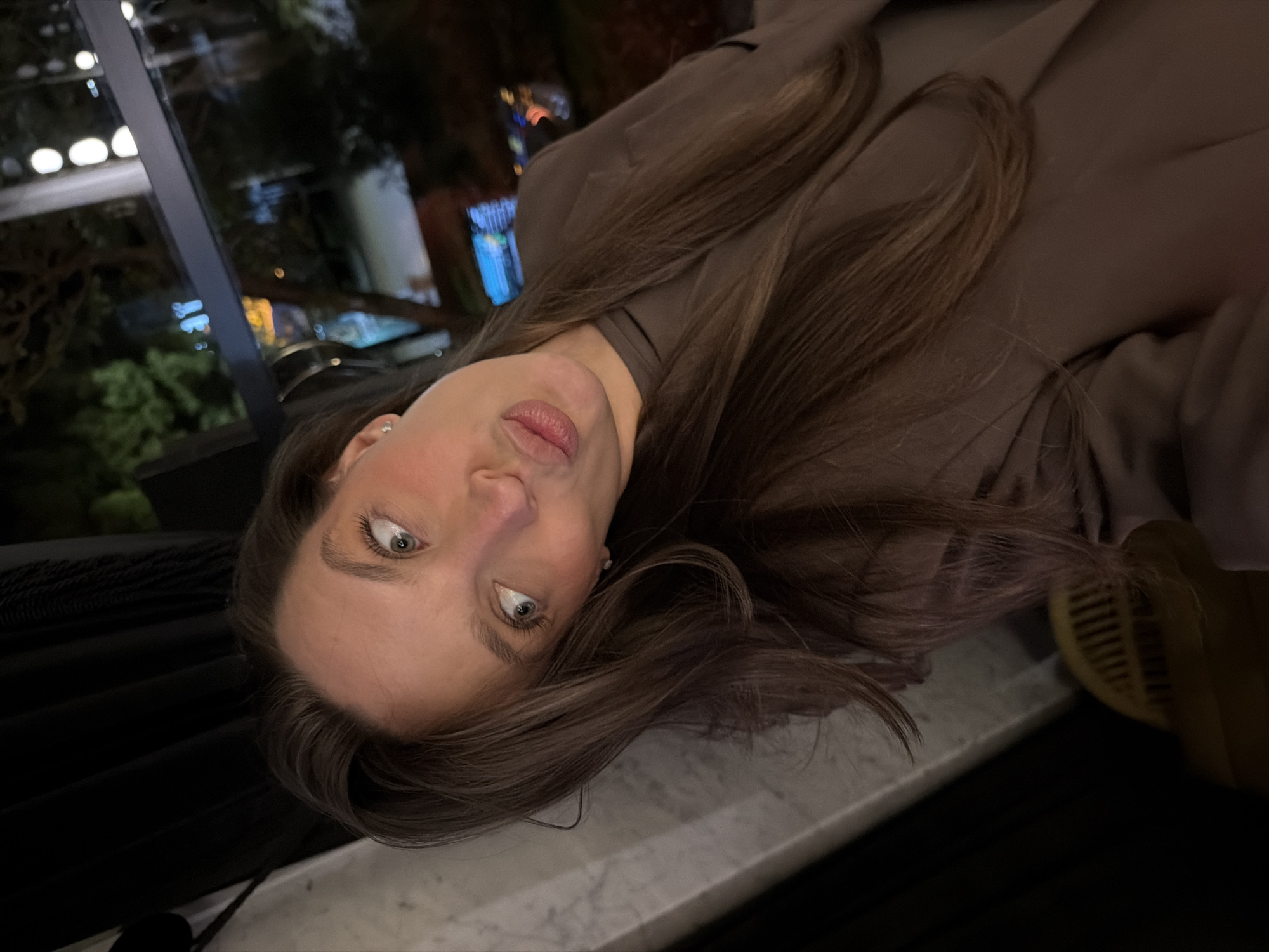 A woman with long brown hair and blue eyes is posing for a close-up selfie. She is wearing a brown blazer or suit jacket. The background shows a blurry view of city lights through a window, suggesting an indoor setting at night. The scene is underexposed. The location cannot be verified to be Budva, Montenegro.