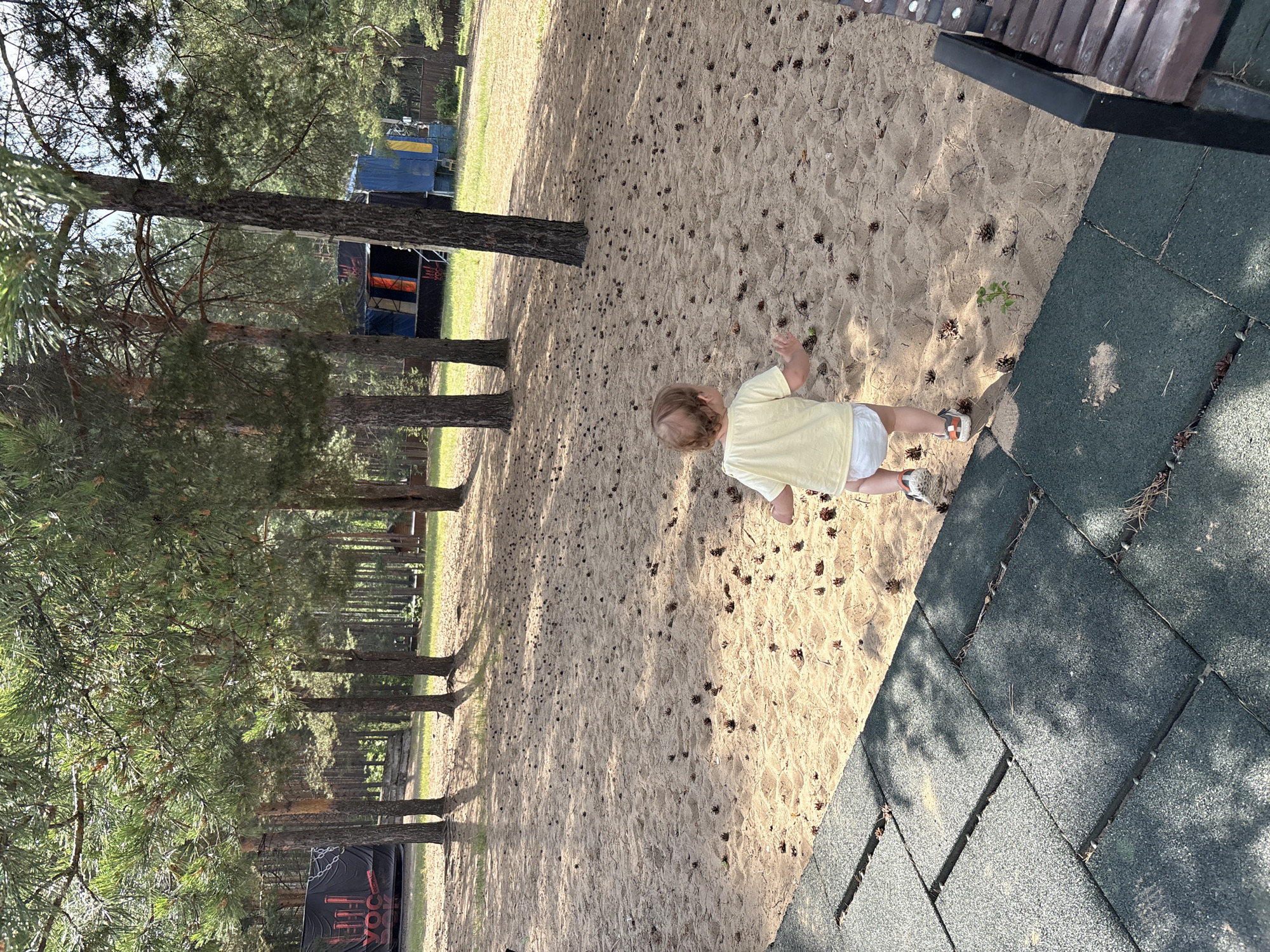 A young child, from the back, wearing a light yellow t-shirt and a diaper, walks on a sandy ground filled with pine cones. The child is on a playground, with dark rubber matting visible in the foreground, and a wooden bench on the right. In the background, there are tall pine trees and a stage with blue and orange curtains. This scene is located in the city of Chaykovsky, Russia.