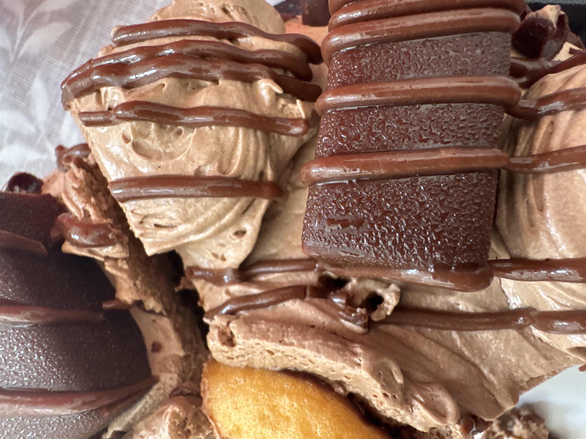 The image is a close-up, overhead shot of a chocolate dessert. The dessert appears to be a cake or layered confection, featuring swirls of chocolate mousse or frosting. Drizzled across the top are dark chocolate sauce or ganache. There are also pieces of what appear to be chocolate bars or truffles, with a ridged texture, interspersed within the dessert. A small, golden-brown, oval-shaped pastry, possibly a madeleine, is visible on the left side of the frame. The lighting is soft, highlighting the glossy texture of the chocolate. The background is out of focus, appearing to be a light-colored, textured surface, possibly fabric or paper. There are no people or any indication of a specific location in Katowice, Poland.