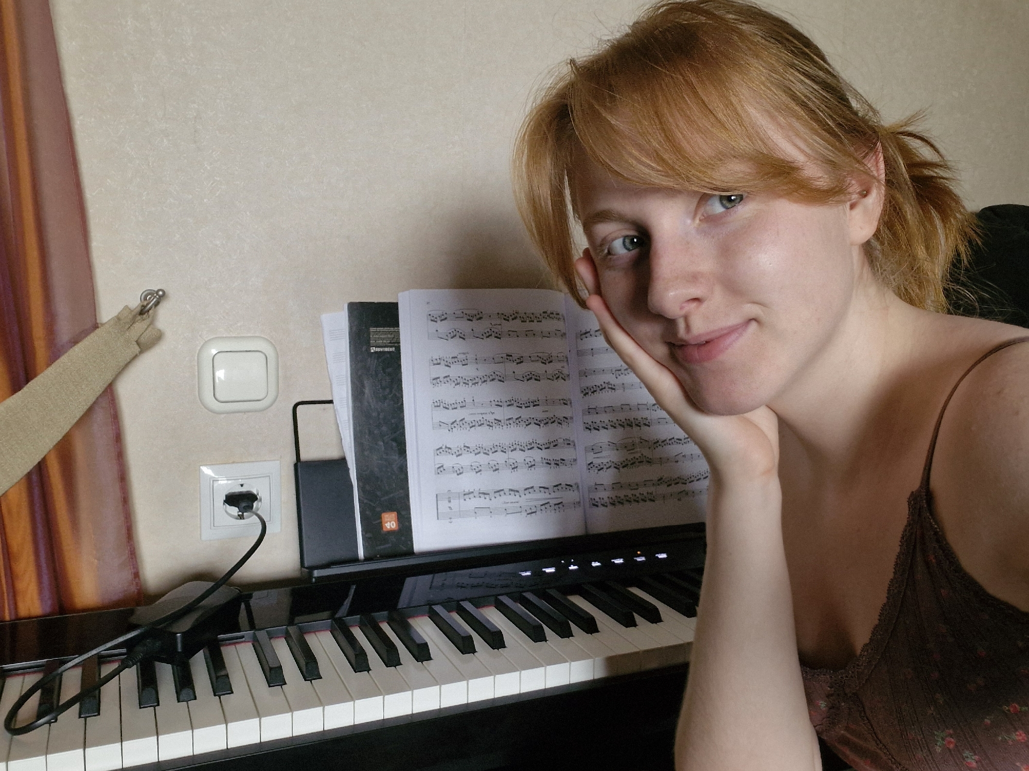 The image features a woman with red hair, styled in low ponytails, resting her face on her hand and gazing at the camera with a slight smile. She is positioned to the right of a black digital piano, which has sheet music displayed on its music stand. A black power adapter is plugged into an electrical outlet above the piano keys. A cream-colored curtain is visible on the left side of the frame, next to a light switch. The background is a plain, light-colored wall. The location and city are unknown.