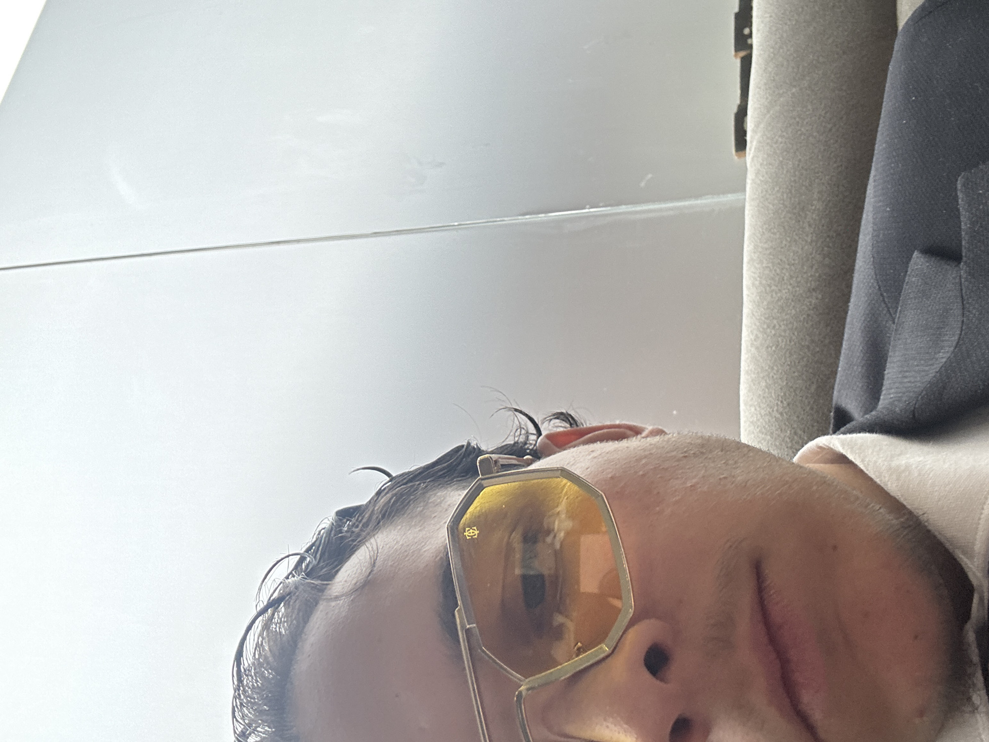 A selfie taken from a low angle shows a person wearing octagonal gold-framed sunglasses. The person's face is partially visible, including their nose, upper lip, and part of their left eye obscured by the sunglasses. The person has dark, wet hair. In the background, there is a white, possibly reflective surface above the person and a grey, textured surface along with a dark-colored jacket to the right. There is no indication of the city of Sofia, Bulgaria, in the image.