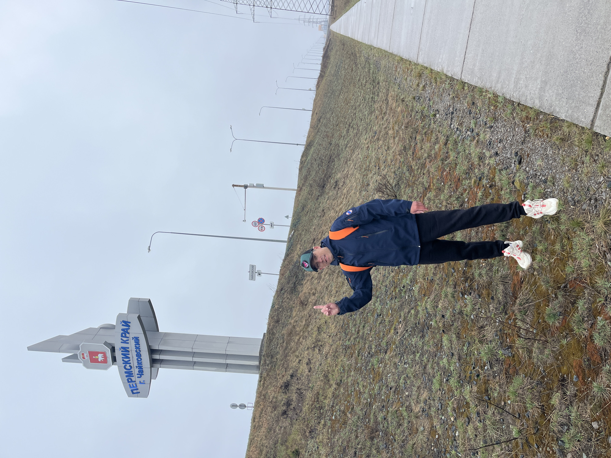 The image captures an outdoor scene on an overcast day, likely alongside a road or highway. In the foreground, a man stands on a sparsely vegetated grassy slope. He is wearing a dark blue jacket with orange shoulder accents, dark trousers, and white and red athletic shoes. He faces the camera, sporting a dark cap and pointing his right index finger slightly downwards.
Behind the man and to his left stands a large, grey, modern-style monument or signpost. The sign is primarily grey with geometric elements and displays text in blue, along with a coat of arms. To the man's right, a concrete path runs parallel to the grassy slope, extending into the distance. In the background, a series of streetlights line the slope, and power lines or pylons are visible further in the distance against the dull, grey sky. Partially visible road signs, including one with a "60" speed limit, are also present.
The text on the monument clearly indicates "ПЕРМСКИЙ КРАЙ" (Permskiy Kray, meaning Perm Krai), and below it, "г. ЧАЙКОВСКИЙ" (g. Chaykovskiy, meaning Chaykovsky city). Another sign, partially obscured, also reads "ЧАЙКОВСКИЙ". This suggests the location is at the entrance or boundary of Chaykovsky city within the Perm Krai region of Russia, consistent with the provided location context of Noviy, Russia, which is in Perm Krai. The time of day is daytime, but the heavy cloud cover gives the scene a muted, cool appearance. The man's pose indicates he is having his picture taken at this landmark.