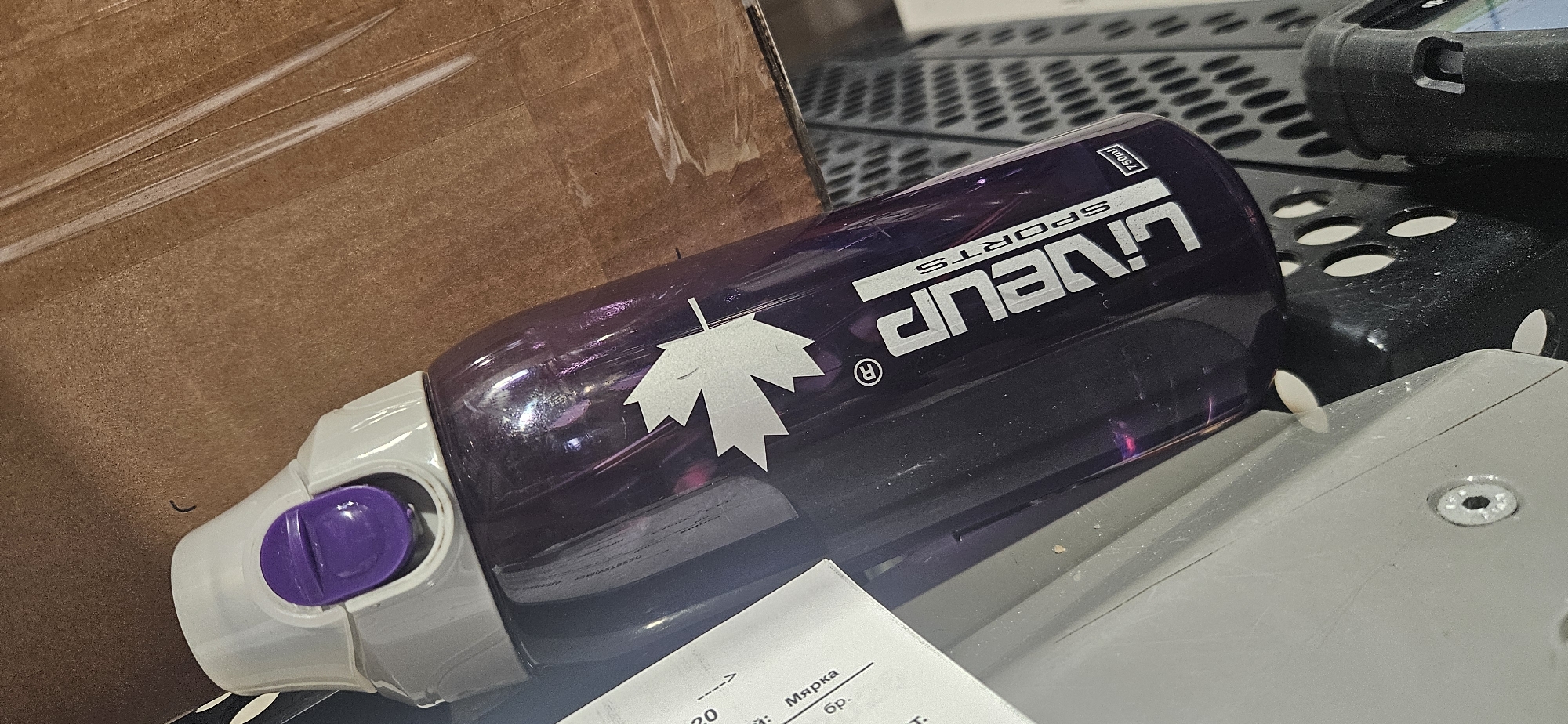 A partially visible purple water bottle with a white maple leaf and the brand name "LINEUP SPORTS" in white lettering lies diagonally across a cluttered surface. A white and purple lid is visible at one end of the bottle. The surface includes a cardboard box, a perforated black surface, a piece of paper with text in Cyrillic, and a gray metal surface with a bolt. The scene is indoors under artificial lighting. The location and city are not discernible from the image.