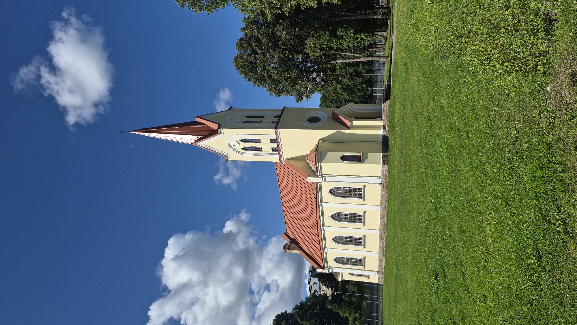 This is a vertical, eye-level, full shot of the Krāslava Evangelical Lutheran Church in Krāslava, Latvia, on a clear, sunny day with scattered white clouds. The church is yellow with a red roof and a tall, slender steeple topped with a cross. The steeple is conical with a red roof and a yellow facade. The church has multiple arched windows along its side. In front of the church is a well-maintained green lawn. To the right of the church, a cluster of mature trees with dark green foliage are visible. In the background, beyond the trees, there is a glimpse of a tennis court and other buildings. The sky is a vibrant blue. There are no people visible in the image.