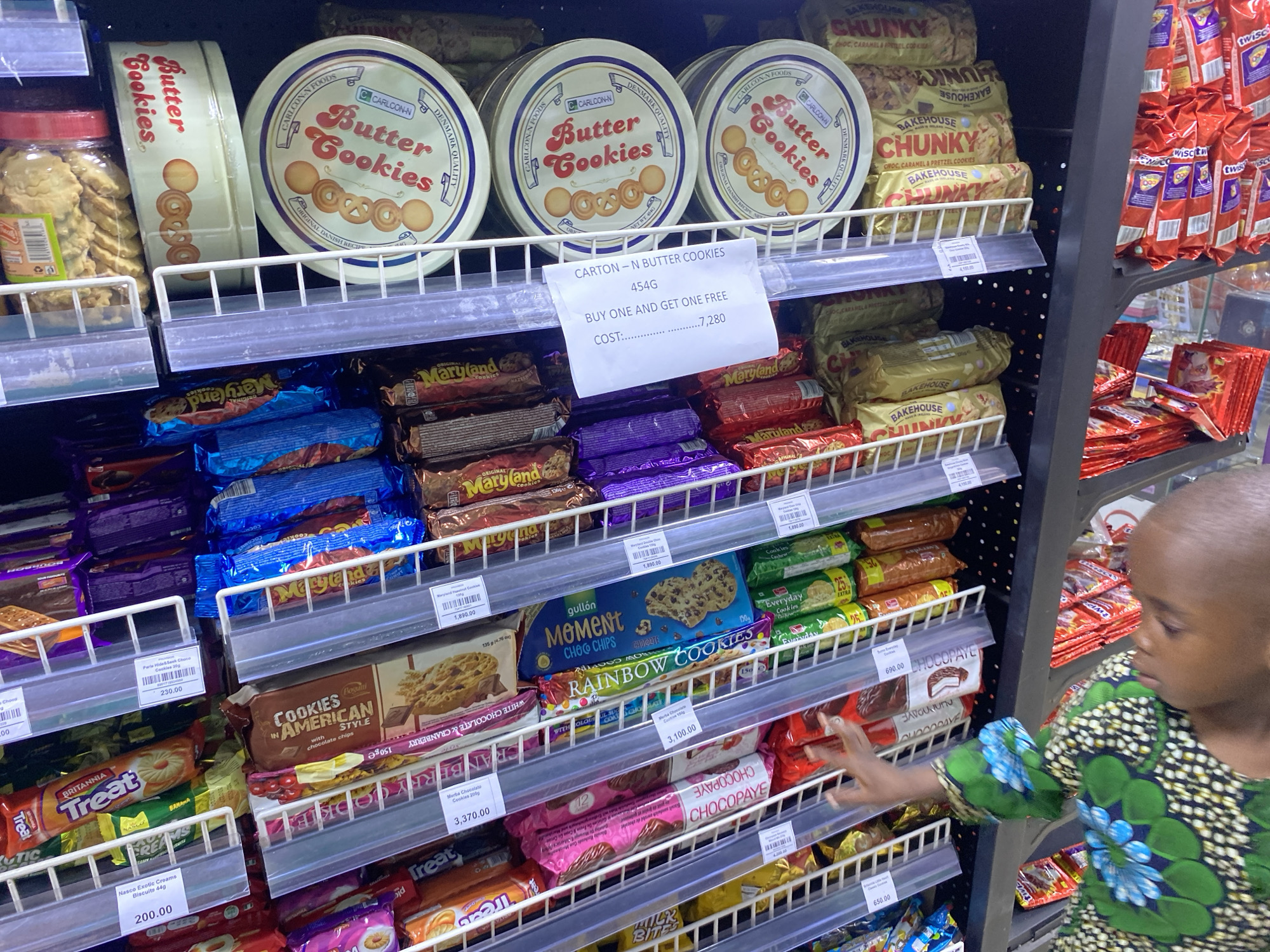 The image shows a retail shelf stocked with various brands of cookies and biscuits in what appears to be a supermarket aisle in Lagos, Nigeria. A young child with a dark complexion and close-cropped hair is reaching towards the cookies on the shelves. The child is wearing a patterned dress with blue and green floral designs.
The shelves display multiple brands including: Carlcon-N Butter Cookies (with a promotion: "Buy one and get one free"), Maryland cookies, Bakehouse Chunky cookies, Rainbow Cookies and Britannia Treat. Price tags are visible for most items. The shelves are white, metal wire racks. The aisle is well-lit.