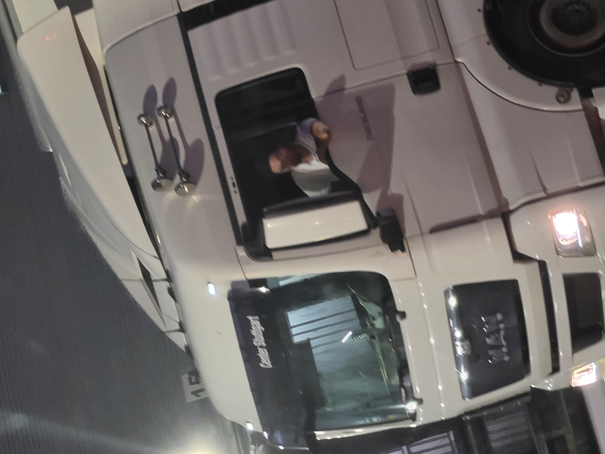 A white semi-truck is parked in what appears to be a commercial area at night. The truck has a dark visor above the windshield with the text "Center Stuttgart" written on it. A man wearing a light-colored shirt is visible through the driver's side window, leaning his elbow on the window frame. The truck's headlights are on, illuminating the immediate surroundings. The background is dark and out of focus, suggesting a nocturnal setting. There is a number "15" visible on a sign attached to the top of the truck. The overall impression is a still shot of a truck and its driver, possibly at a loading dock or a service area.