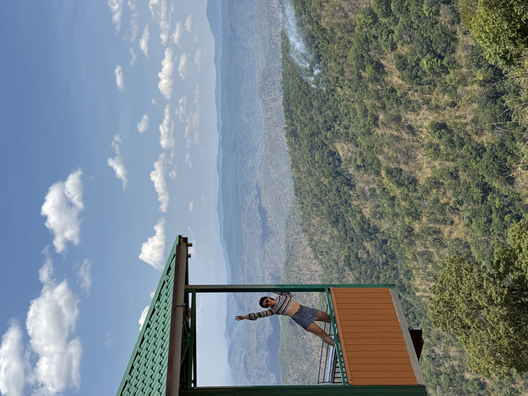 The image captures a vibrant mountainous landscape with a person posing on a viewing platform. The scene is dominated by layers of green mountains stretching into the distance under a bright blue sky dotted with scattered white clouds. The mountains are covered in dense forest, with some patches appearing drier or less vegetated. A plume of white smoke, possibly from a small fire, rises from the trees on one of the closer hills.
In the foreground, a person, appearing to be a young woman, is standing on a balcony or observation deck. She is wearing a striped long-sleeved crop top and denim shorts. She is leaning backward, extending her arms out to her sides, striking a playful or energetic pose as she takes in the expansive view. The structure she is standing on features a green corrugated roof visible on the left, and a wooden-paneled section on the right, framing the mountainous panorama. A dark metal railing is present at the edge of the platform.
The weather is clear and sunny, indicative of daytime. The lush greenery and layered mountains suggest a scenic, possibly remote, natural location in Phan Son, Vietnam. There is no visible text in the image.