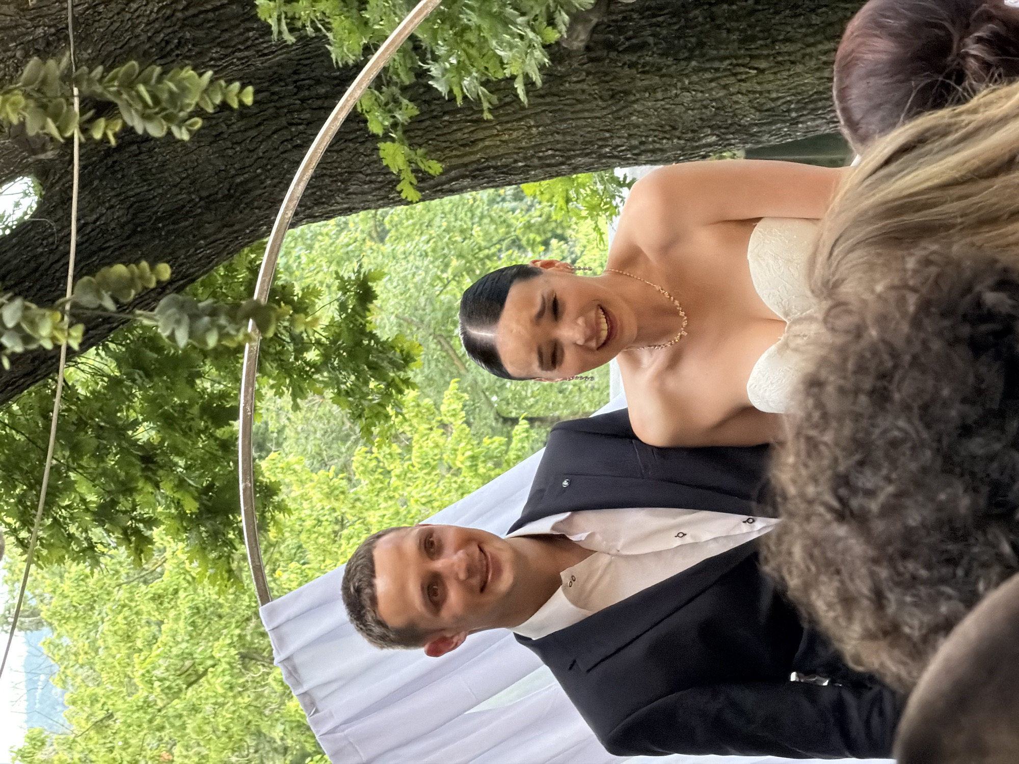 A medium shot captures a man in a suit and a woman in a wedding dress during an outdoor wedding ceremony in Tbilisi, Georgia. The man, on the left, smiles warmly at the camera, dressed in a black suit jacket and a white button-down shirt. The woman, to his right, is laughing, her head tilted back, with her eyes closed and mouth open. She is wearing a strapless white wedding dress and a delicate necklace. They are positioned under a floral archway, with white drapes to their left and a large tree trunk to their right. The background shows lush green foliage and trees, suggesting a natural setting within the city. The focus is on the couple, with the blurred heads of wedding guests visible in the foreground.