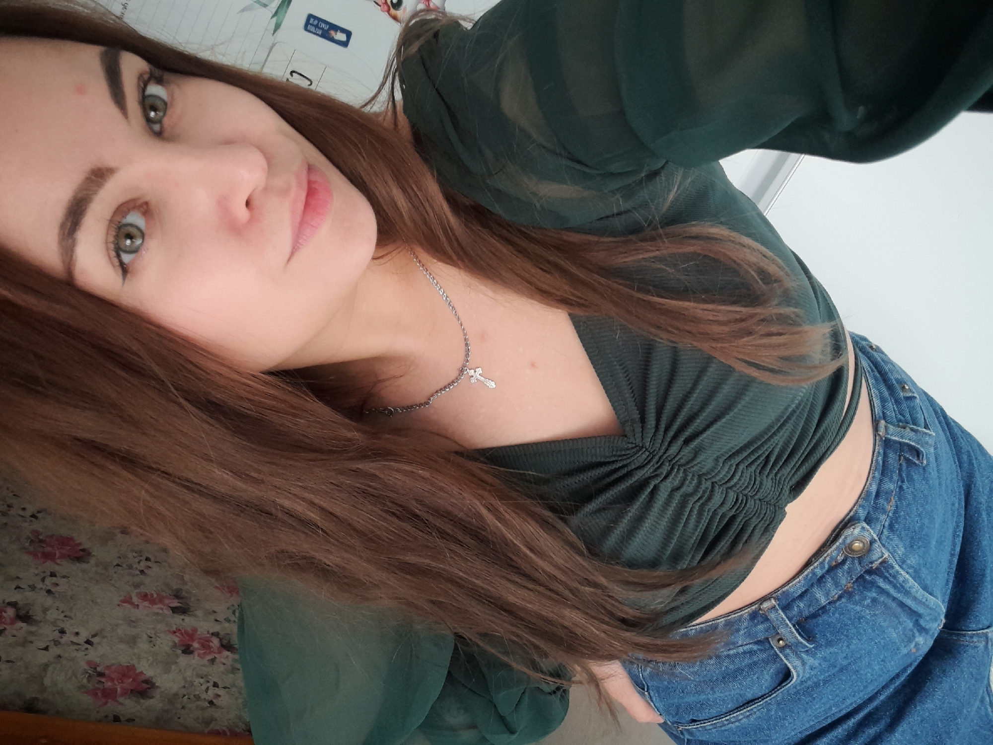 A selfie photograph shows a woman with fair skin, brown hair, and green eyes. She is wearing a dark green, short-sleeved shirt and blue jeans. A silver necklace with a cross pendant is visible around her neck. The background includes a floral pattern on a surface to her left, and a white wall behind her. The photo appears to be taken indoors, with no discernible location in the city of Unknown, Unknown.