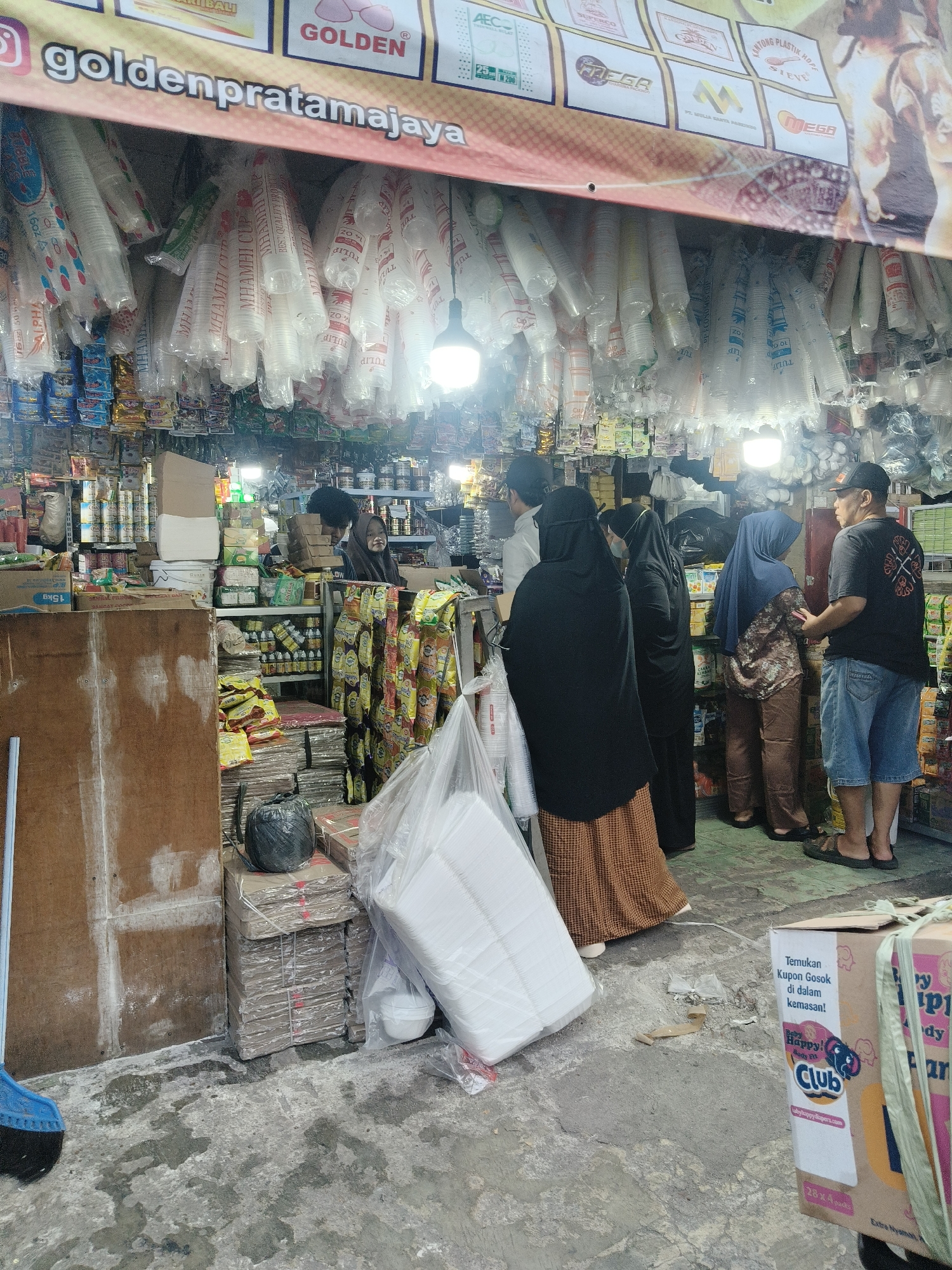 The image shows a street-level stall in South Tangerang, Indonesia, selling various goods. In the foreground, a blue broom leans against a large wooden panel. The floor is concrete, showing signs of dampness and debris. Behind the wooden panel, stacks of cardboard boxes are visible, some bound with string and plastic. A large black garbage bag is placed on top of some of these boxes. To the right, a large cardboard box with "Baby Happy!" branding is prominently displayed.
The stall itself is filled with goods hanging from the ceiling, appearing to be plastic cups packaged in transparent plastic. Shelves behind the counter are stocked with various packaged food items and other retail products. Several people are present in the stall area.
On the left, a woman in a dark hijab and an orange and brown patterned skirt is partially visible. Next to her, another person in a black hijab faces away from the camera. Further to the right, a person wearing a blue hijab and a patterned shirt stands with their back to the viewer. A man in a black t-shirt and denim shorts, wearing a black cap, stands facing the group, engaged in conversation. The background shows more shelves filled with products and lighting fixtures. The overall scene depicts a busy, functional retail environment.