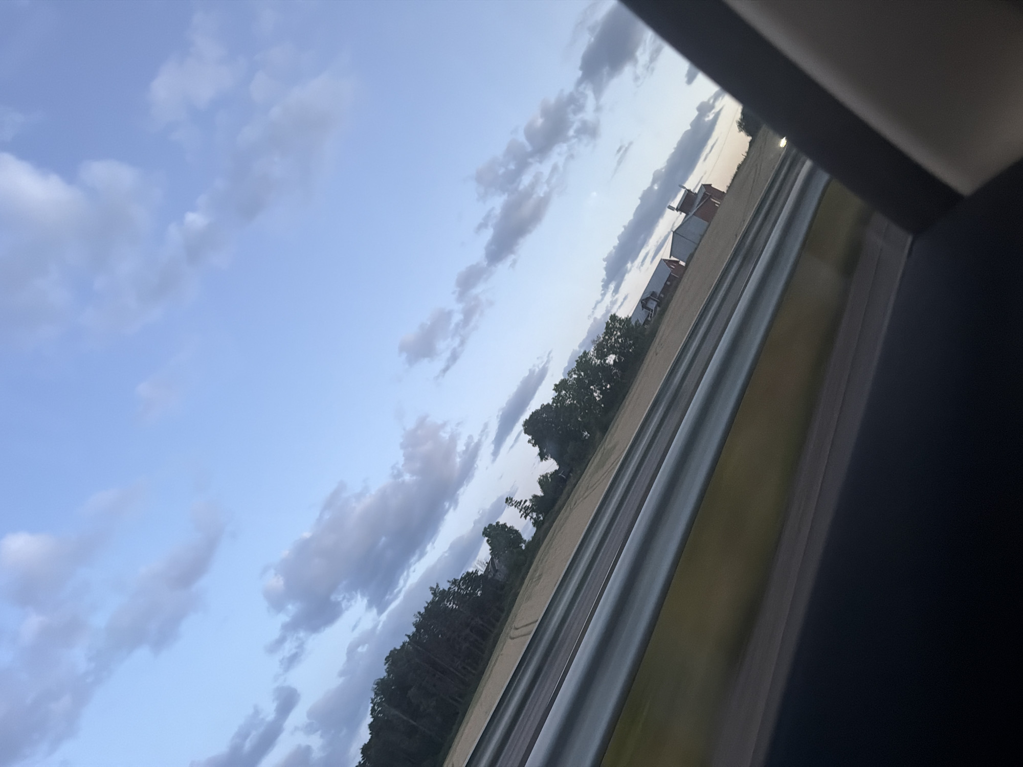 The image shows a rural landscape with a field, trees, and buildings, seen from a vehicle. The sky is partly cloudy. The scene appears to be a country road in Sarpsborg, Norway. There are no people or animals visible. The focus is on the natural scenery and the architecture of the farm buildings.