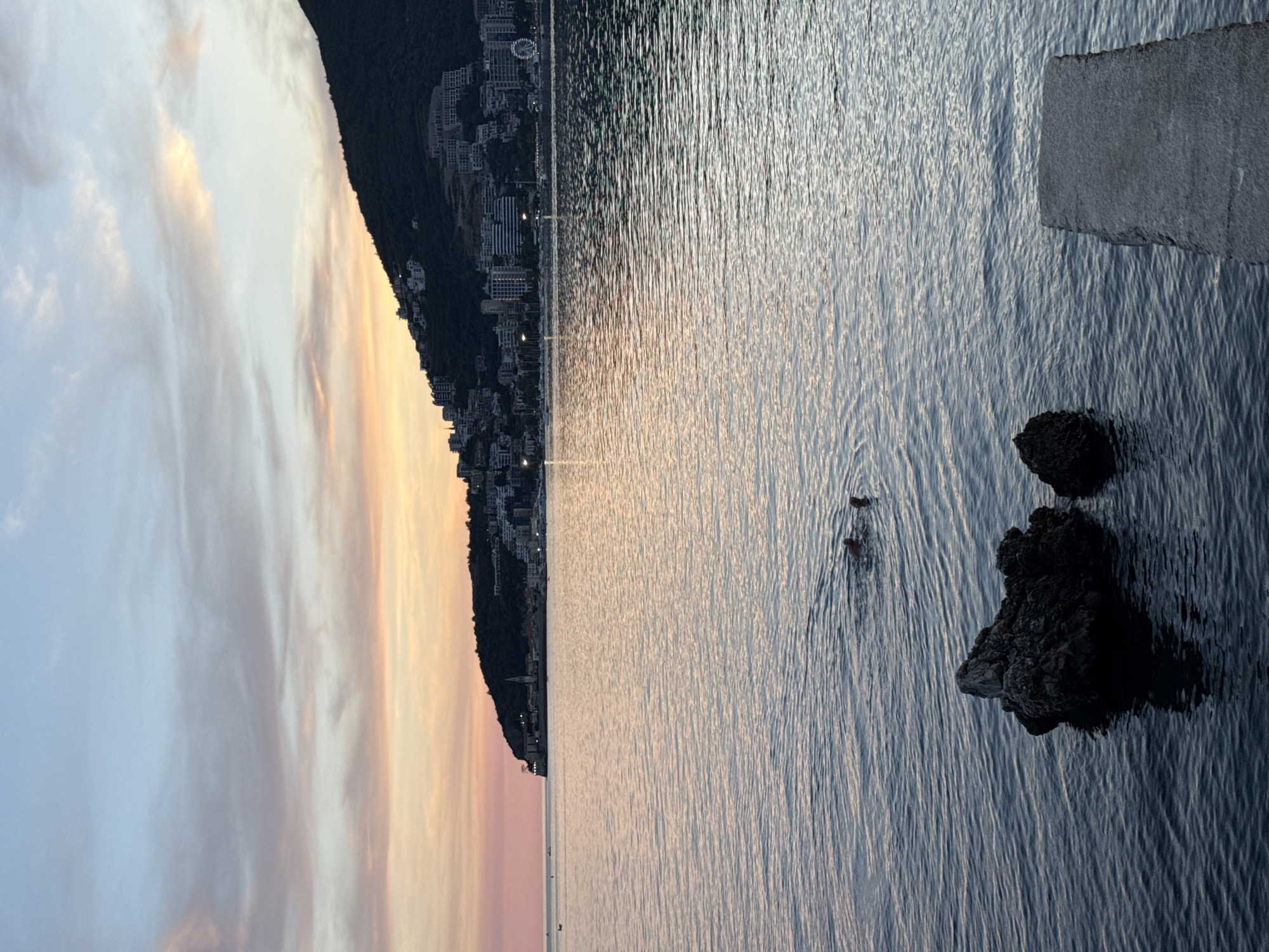 The image depicts a serene sunset over the Adriatic Sea, with the coastline of Budva, Montenegro, forming the backdrop. In the foreground, dark, jagged rocks emerge from the calm, rippling water. Further out, two individuals are swimming, their heads visible above the surface. The water transitions from a deep blue in the foreground to a lighter, shimmering surface reflecting the pastel hues of the sky.
The middle ground is dominated by the expansive sea, its surface gently disturbed by subtle waves. In the distance, the city of Budva stretches along the base of a steep, dark green hill. Buildings, appearing as clusters of white and light-colored structures, cling to the hillside, suggesting a densely populated urban area. Several lights from the city twinkle faintly against the darkening landscape. A ferris wheel, a prominent landmark, is visible on the right side of the city.
The background is a canvas of soft twilight colors. The sky is a gradient of pale blue at the top, transitioning through delicate shades of pink, peach, and orange near the horizon. Wispy clouds are scattered across the sky, catching the last rays of the setting sun and adding a touch of drama to the tranquil scene. The overall impression is one of peacefulness and natural beauty, with human presence subtly integrated into the coastal landscape.