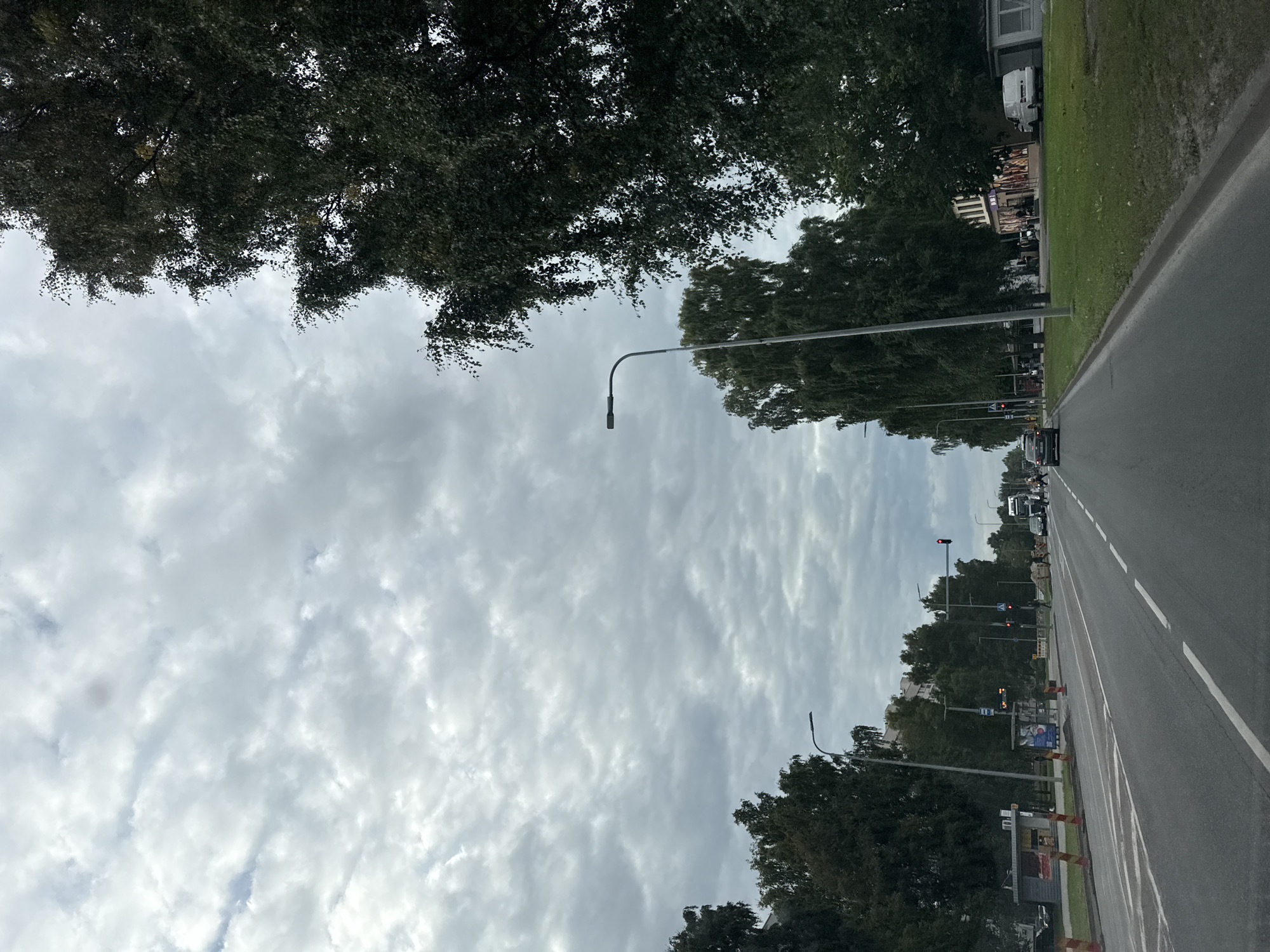 The image depicts a street scene in Tallinn, Estonia, on a cloudy day. The perspective is from a vehicle driving down a multi-lane road. On the left side of the road, there are trees, a bus stop with a shelter, some buildings, and traffic cones indicating construction. On the right side of the road, there are more trees, grass, and buildings with storefronts. Several cars are visible on the road, including a white van parked on the right. Pedestrians can be seen crossing the street in the distance. Traffic lights are visible, with one showing red. The sky is overcast with textured clouds.