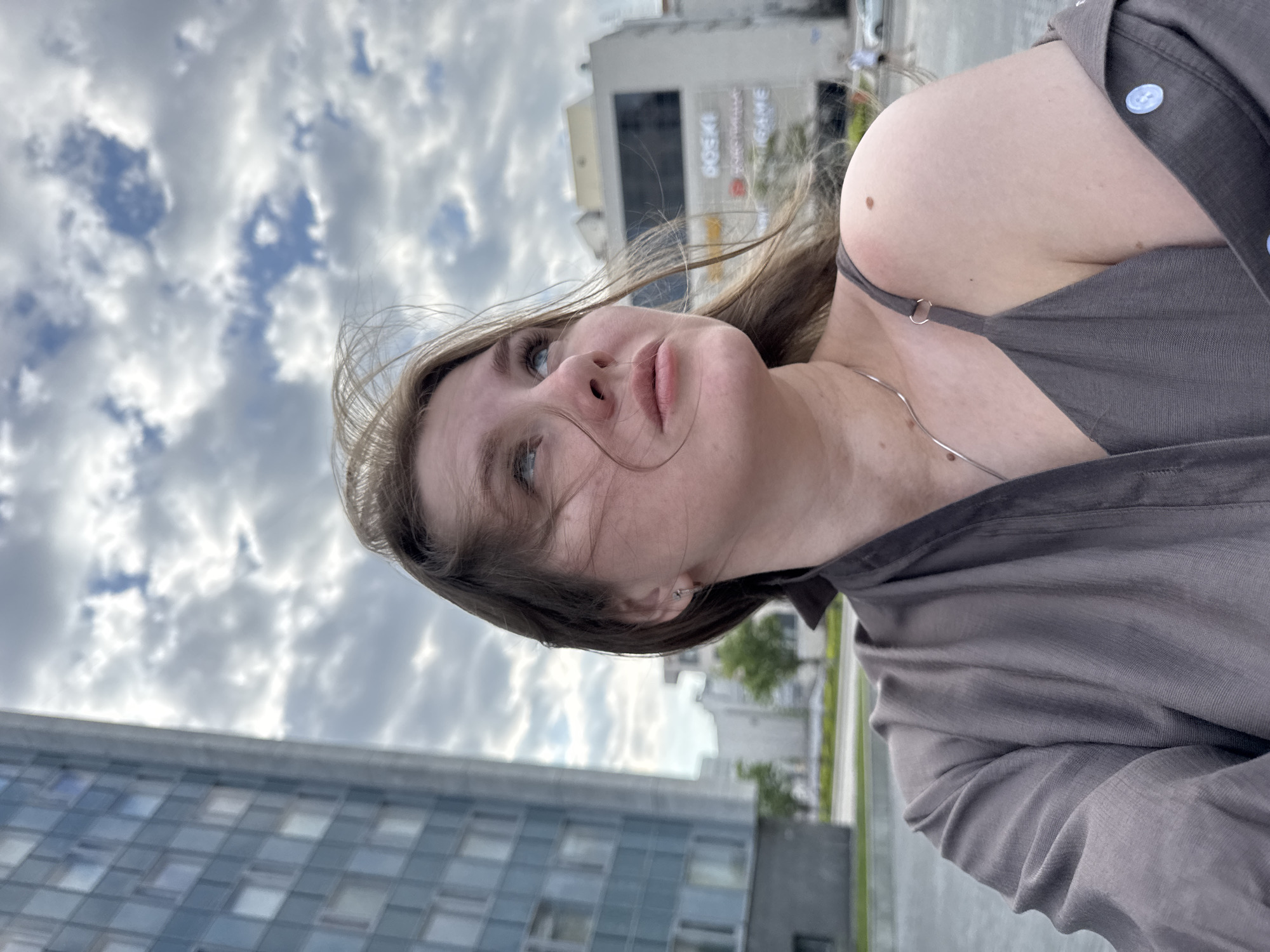 A woman poses in Perm, Russia, looking upward. The sky is partially cloudy. A large, modern building with reflective windows is visible in the background, as is a building with writing in the Russian language. The woman wears a gray top with thin straps and a silver necklace. She has light skin, brown hair, and blue eyes.