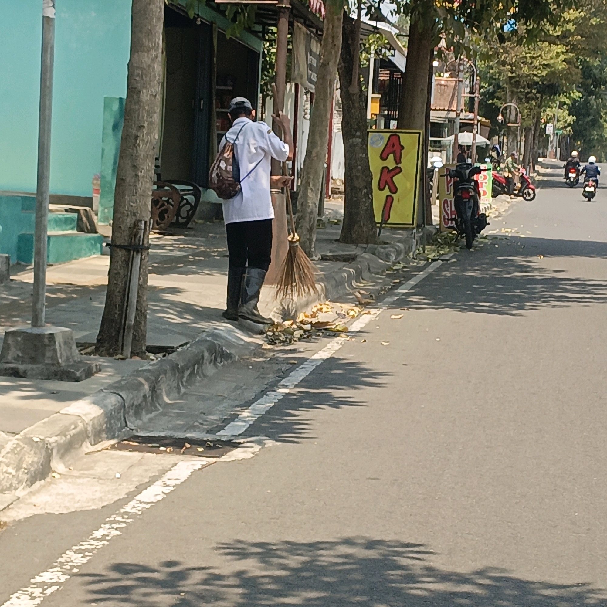 The image depicts a street scene in Salatiga, Indonesia. In the foreground, a person wearing a white shirt, black pants, and black boots is sweeping fallen leaves and debris from the sidewalk into the street with a broom. The person has a patterned backpack slung over their left shoulder. Adjacent to the sidewalk are trees, one of which has a bundle of long sticks tied to it. A yellow sign with red lettering, appearing to spell "AKI," is visible behind the person and to their right, mounted on a pole. Further down the street, several motorcycles are visible, some ridden by people wearing helmets. The street is paved, with a white dashed line marking a lane. The environment appears to be a commercial or residential area with buildings and trees lining the street. The lighting suggests it is daytime, with shadows cast on the road by trees and possibly overhead structures.