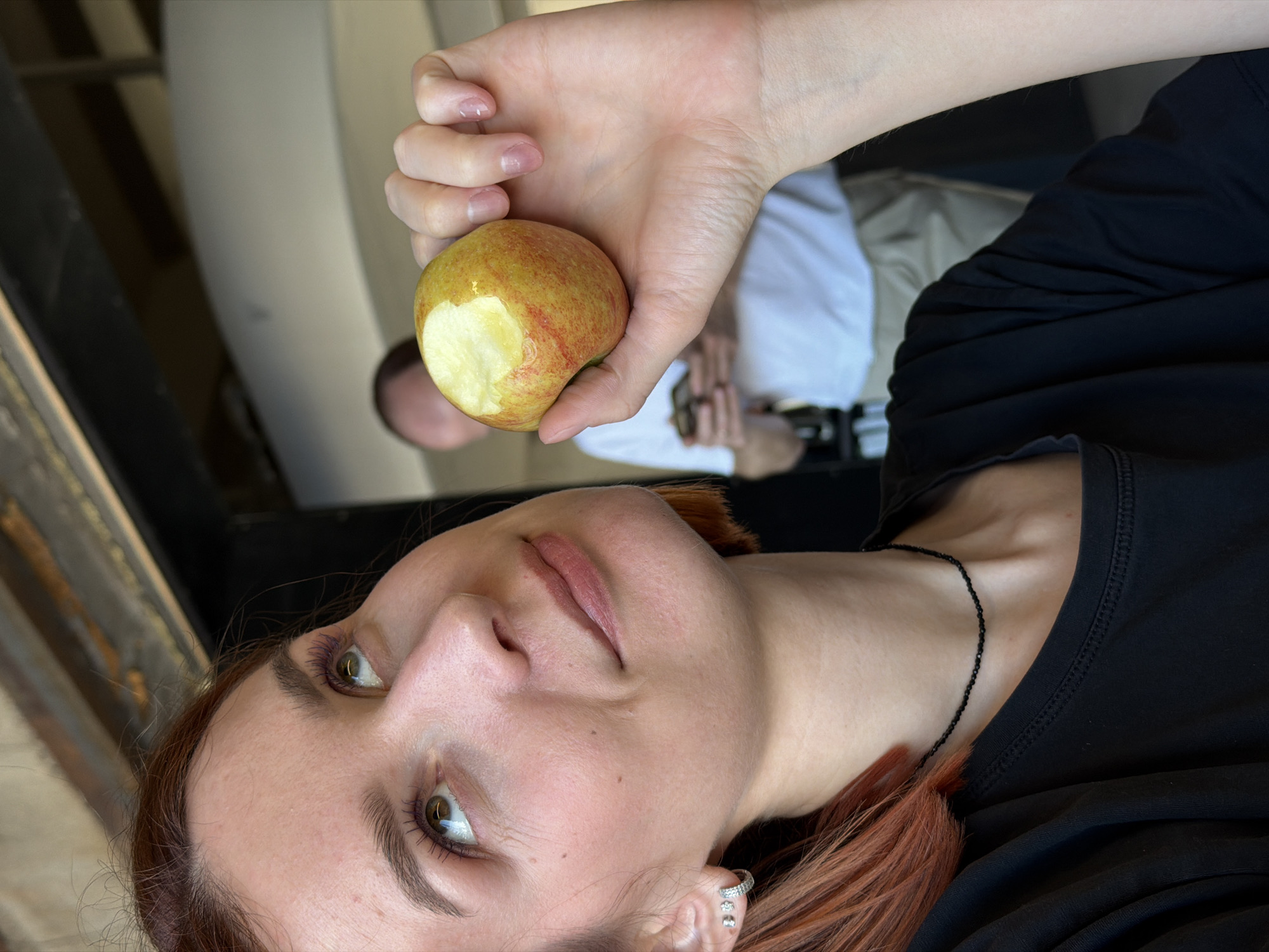 A woman with fair skin and auburn hair is holding a partially eaten apple in her right hand. She is wearing a black top and a delicate black necklace. Her gaze is directed upwards. The background features a blurred figure of a man wearing a white shirt and khaki pants, also holding a camera, seemingly taking a photograph. Metallic structures are partially visible, indicating an urban setting, possibly in Chaikovsky, Russia.