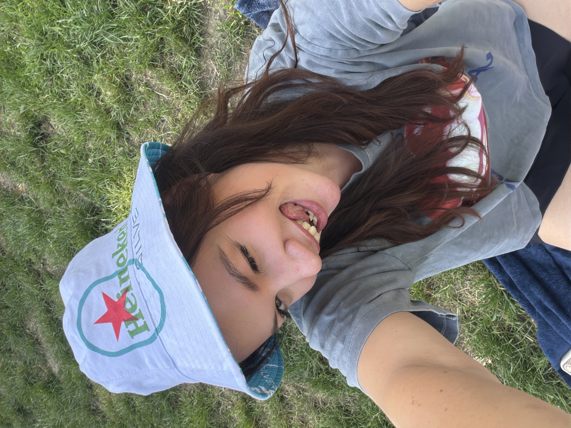 A young person lies on a grassy surface in Cukarica Urban Municipality, Serbia. They are wearing a light-colored bucket hat with a Heineken logo, a gray t-shirt, and dark shorts. The person's hair is dark brown, and they are sticking out their tongue. A blue towel is near them. The ground is covered in green grass.