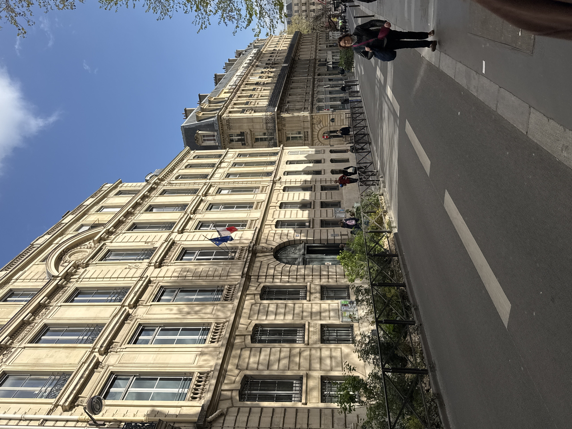The image shows a street in Paris, France, with buildings displaying classical Parisian architecture. The buildings are several stories high with numerous windows. French flags are visible on one of the buildings. There are people walking on the sidewalk alongside the road. A black metal fence lines the sidewalk. The road is paved with asphalt, and lane markings are visible. The sky is clear and blue with some clouds. The photograph appears to have been taken during the day.