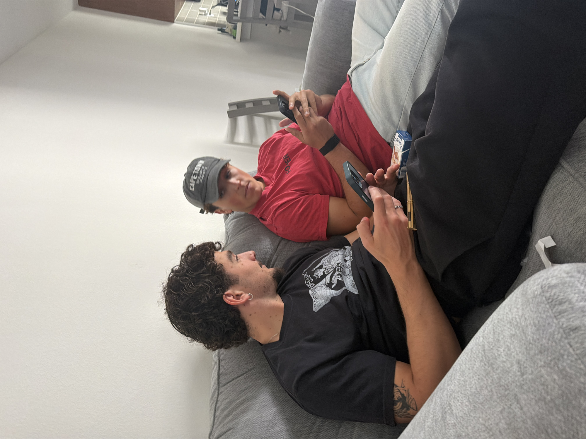Two young men are shown lounging on a grey upholstered couch in a bright, minimalist indoor setting, likely a living room, in Redondo Beach, United States of America. The room features stark white walls and clear, even lighting, suggesting it is daytime.
The man in the foreground, with dark, curly hair and a short beard, is wearing a black short-sleeved t-shirt with a white graphic print. He has silver hoop earrings, a silver chain, and a visible tattoo on his left forearm. He holds a black smartphone in his right hand, looking up with a neutral or slightly curious expression, possibly at the other man or his phone.
Behind him is the second man, wearing a red short-sleeved shirt and a grey baseball cap that clearly displays "CAPE TOWN" on the front. A black smartwatch or fitness tracker is on his left wrist. He is holding a smartphone with both hands, seemingly engrossed in its screen. A pack of "CAMEL" brand cigarettes is visible tucked into the couch cushions beside him.
Both individuals appear relaxed and are engaged with their personal electronic devices, illustrating a common contemporary scene of casual digital interaction. The overall atmosphere is one of leisure and comfort in a modern domestic environment.