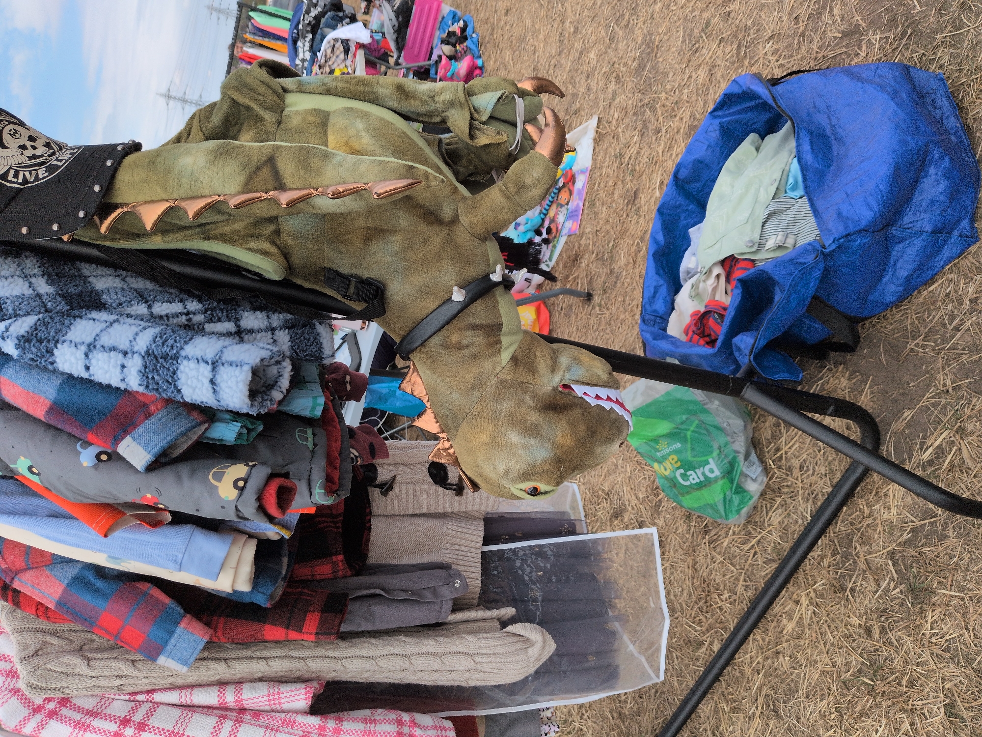 This image shows a close-up of a rack of children's clothing displayed at an outdoor market. In the foreground, a large stuffed dinosaur costume is hanging on the rack, with its head visible. The dinosaur is green with bronze-colored spikes down its back and has its mouth open, revealing red and white teeth. It is wearing a black hat with a skull emblem. A black harness is around the dinosaur's neck.
To the left of the dinosaur, several items of clothing are hanging on the rack, including patterned shirts and sweaters. A clear plastic garment bag hangs below some of the clothes. A green plastic shopping bag with the word "Morrisons" is on the ground to the left of the rack.
In the lower right of the image, a large open blue bag is filled with folded clothes. The background shows a dry, grassy ground, typical of an outdoor setting. Further back, other market stalls with various items are visible, suggesting this is a flea market or car boot sale. The sky above is overcast. The scene is consistent with an outdoor market setting within the city of South Staffordshire, United Kingdom, likely during the day given the lighting. There are no people visible in this particular frame.