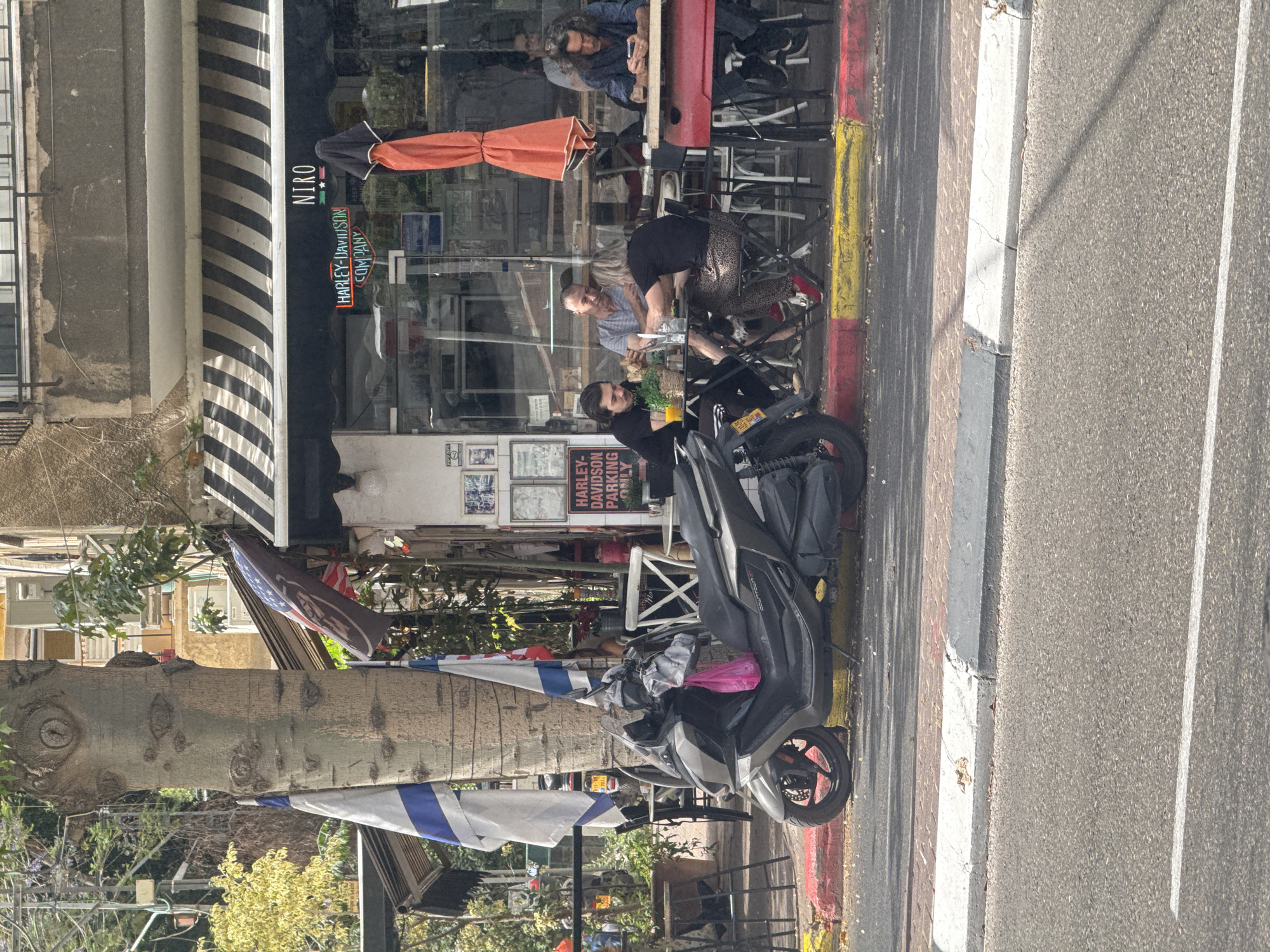 The image shows a street scene in Givatayim, Israel, featuring the "NIRO" establishment, possibly a cafe or restaurant, where several individuals are seated at outdoor tables. A black scooter is parked on the sidewalk in front of the establishment, adorned with what appears to be personal belongings. A sign indicating "HARLEY DAVIDSON PARKING ONLY" is visible near the scooter. An Israeli flag is prominent alongside the scooter. The image captures a casual daytime scene in an urban setting.