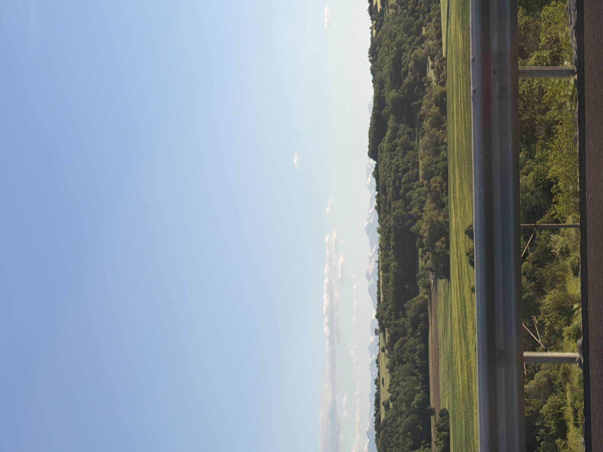 The image depicts a landscape view in Vakarel, Bulgaria. The sky is predominantly clear and blue. To the right, there is a lush green landscape consisting of trees, meadows, and what appears to be cultivated fields. A roadside barrier is present on the right side of the image, giving the impression of looking out from a roadside. In the distance, faintly visible, are possibly snow-capped mountain peaks.