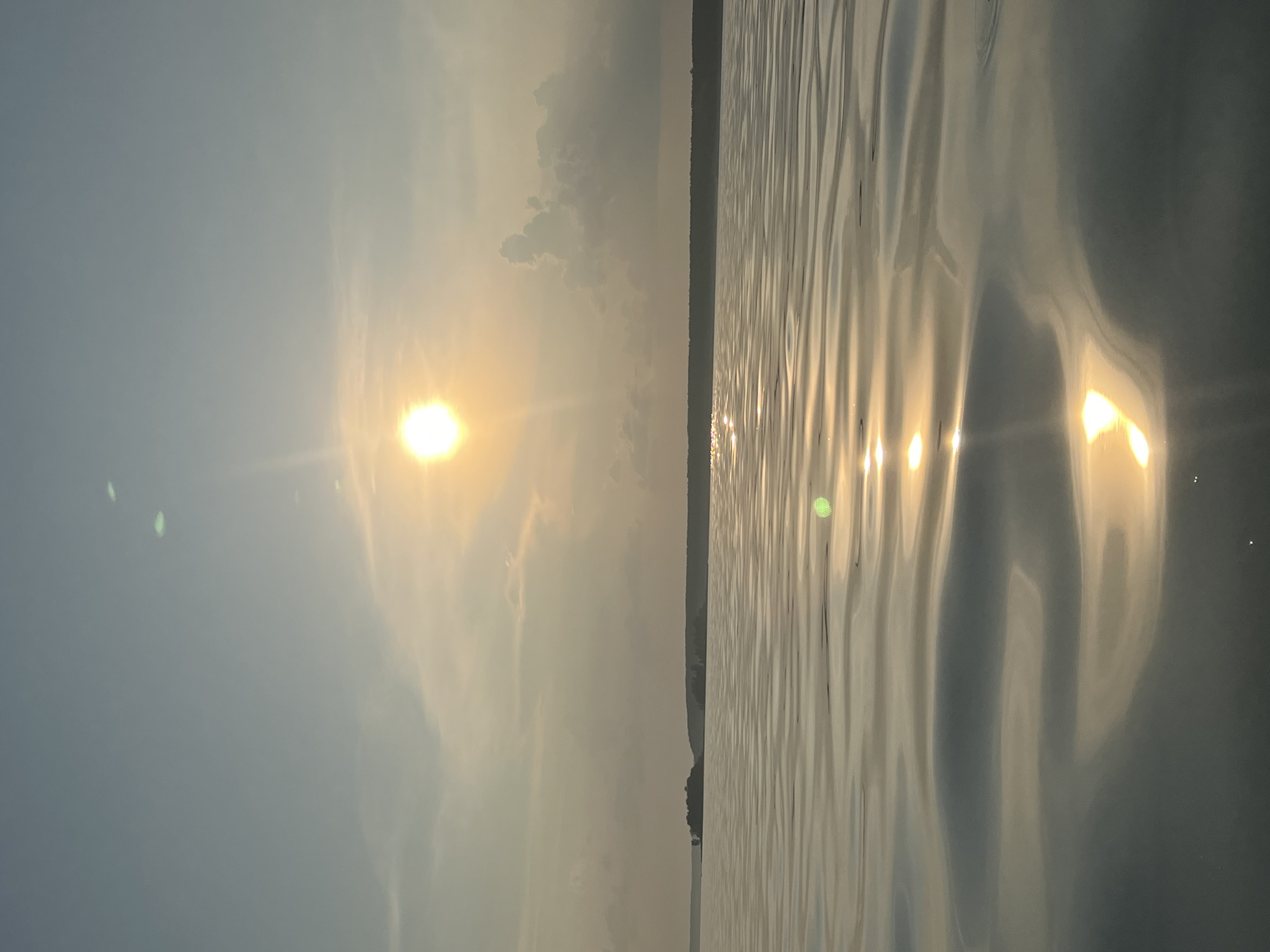 The image depicts a serene body of water, likely a lake, under a hazy sky with the sun visible as a bright, diffused orb near the center. The water's surface is rippled, reflecting the sunlight in shimmering patterns. In the distance, a dark, treeline forms the horizon, with a small, isolated island visible to the left, also covered in vegetation. The overall atmosphere is calm and natural, suggesting an outdoor scene at a natural reserve or park. The location provided, Borovoe, Russia, is consistent with a landscape featuring a large lake and forested areas. No people or specific human activities are evident in the image.