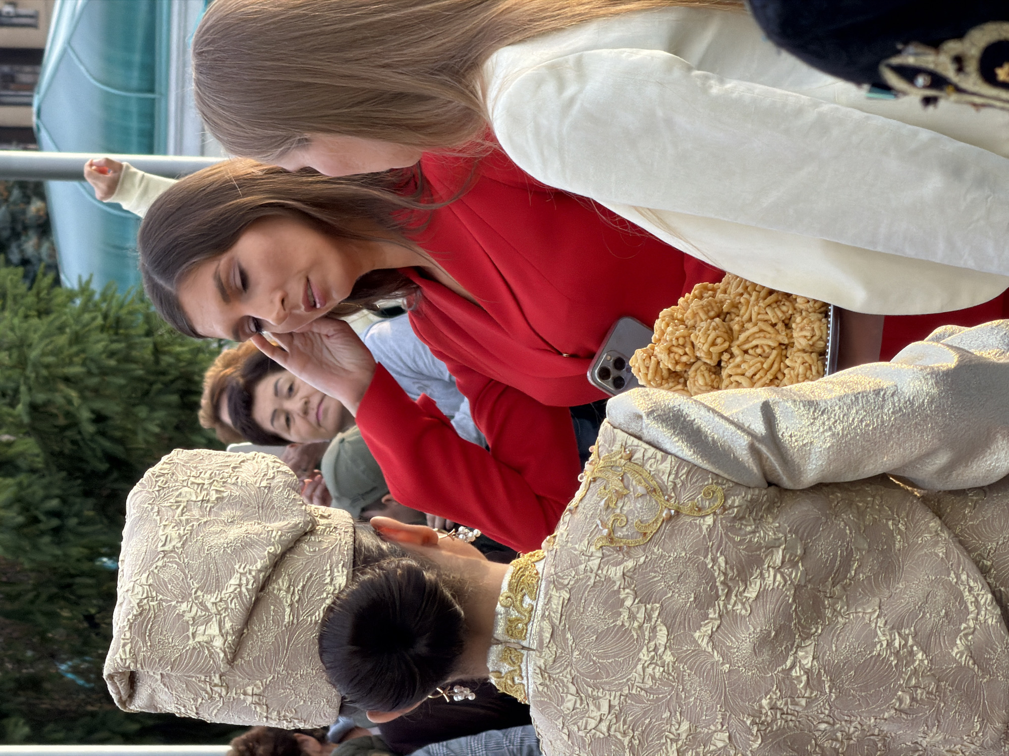 A group of people are gathered outdoors, likely in a public square in Unknown, Unknown, as indicated by the presence of a large building with a green roof in the background and trees. In the foreground, three women are the primary focus. The woman on the left is seen from the back, wearing an elaborate gold and cream patterned outfit with a matching head covering and dangling pearl earrings. She has her dark hair pulled back into a bun. Next to her, a woman in a bright red blazer is turned towards the woman on the far right, her left hand touching her temple. She has long, brown hair and appears to be speaking or listening intently. The woman on the right is facing away from the camera, her right side visible. She has long, blonde hair and is wearing a cream-colored blazer. Between the woman in red and the woman on the right, a tray of chak-chak, a traditional Tatar pastry made of fried dough balls coated in honey, is visible. A mobile phone is also resting on the tray. Other people are partially visible in the background, suggesting a social gathering or event.