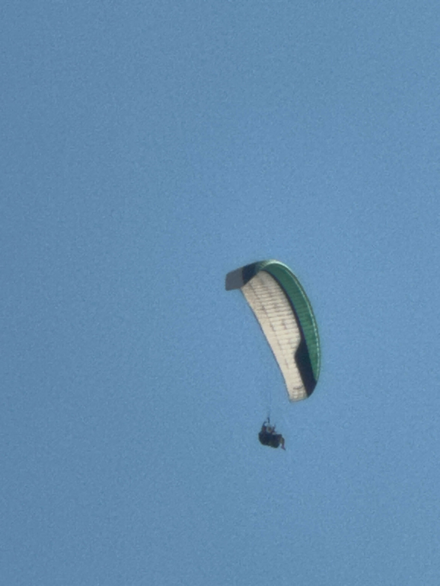 A paraglider, its canopy a mix of white, green, and black, is captured in mid-air against a clear blue sky. A person is seated in the harness suspended beneath the paraglider. The paraglider appears to be in a slightly tilted position. The image does not contain any identifiable landmarks or features that would allow for the determination of its location within Boreti, Montenegro.