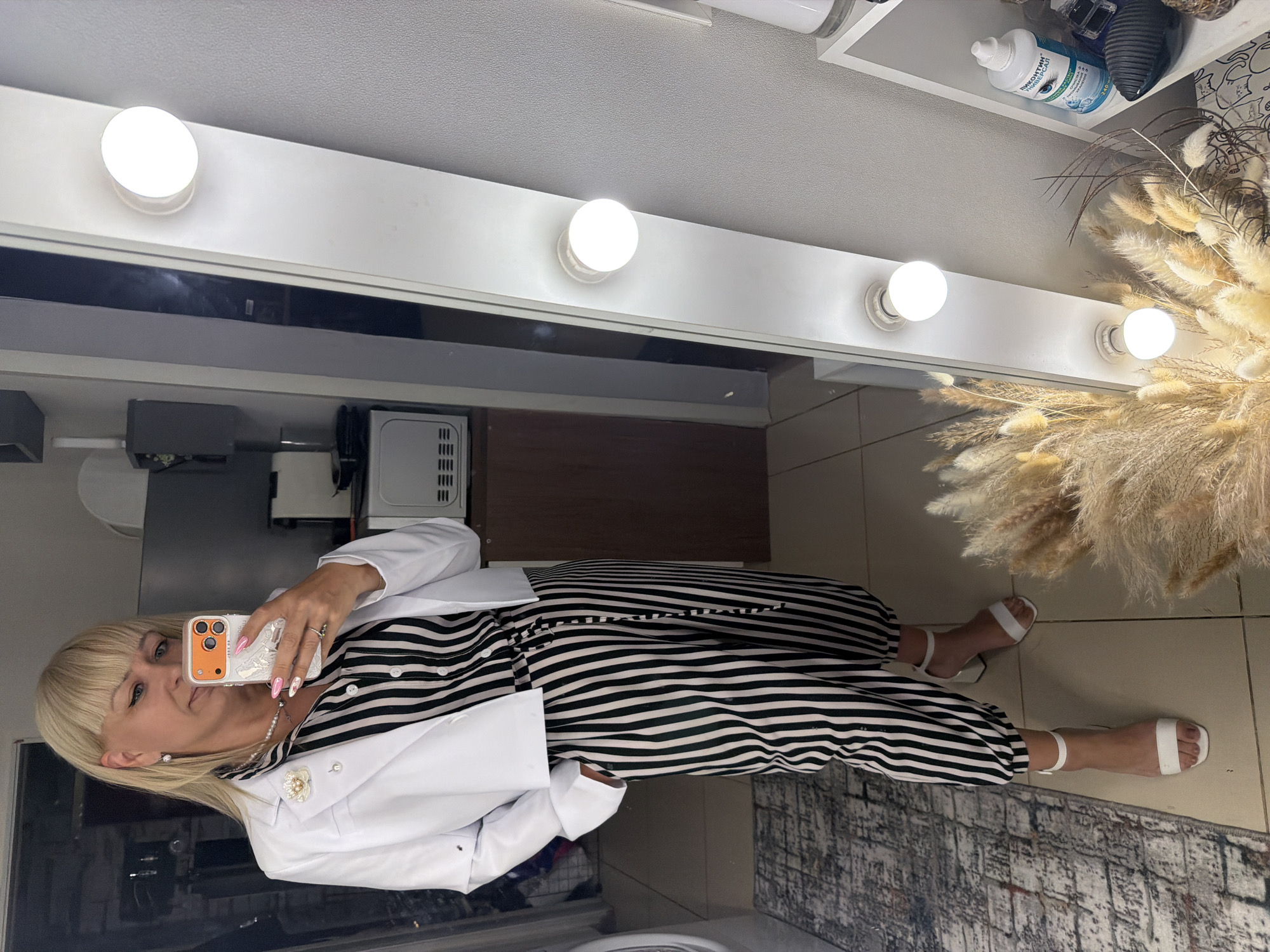 A middle-aged woman with blonde bangs is taking a selfie in what appears to be a bathroom or dressing room. She is wearing a striped jumpsuit with a white blazer. The walls are tiled and there is a rug on the floor. In the background, there is a vanity mirror with light bulbs and some decorative items. Without metadata or additional information, pinpointing the location to Chaikovsky, Russia, cannot be forensically verified from the imagery alone.