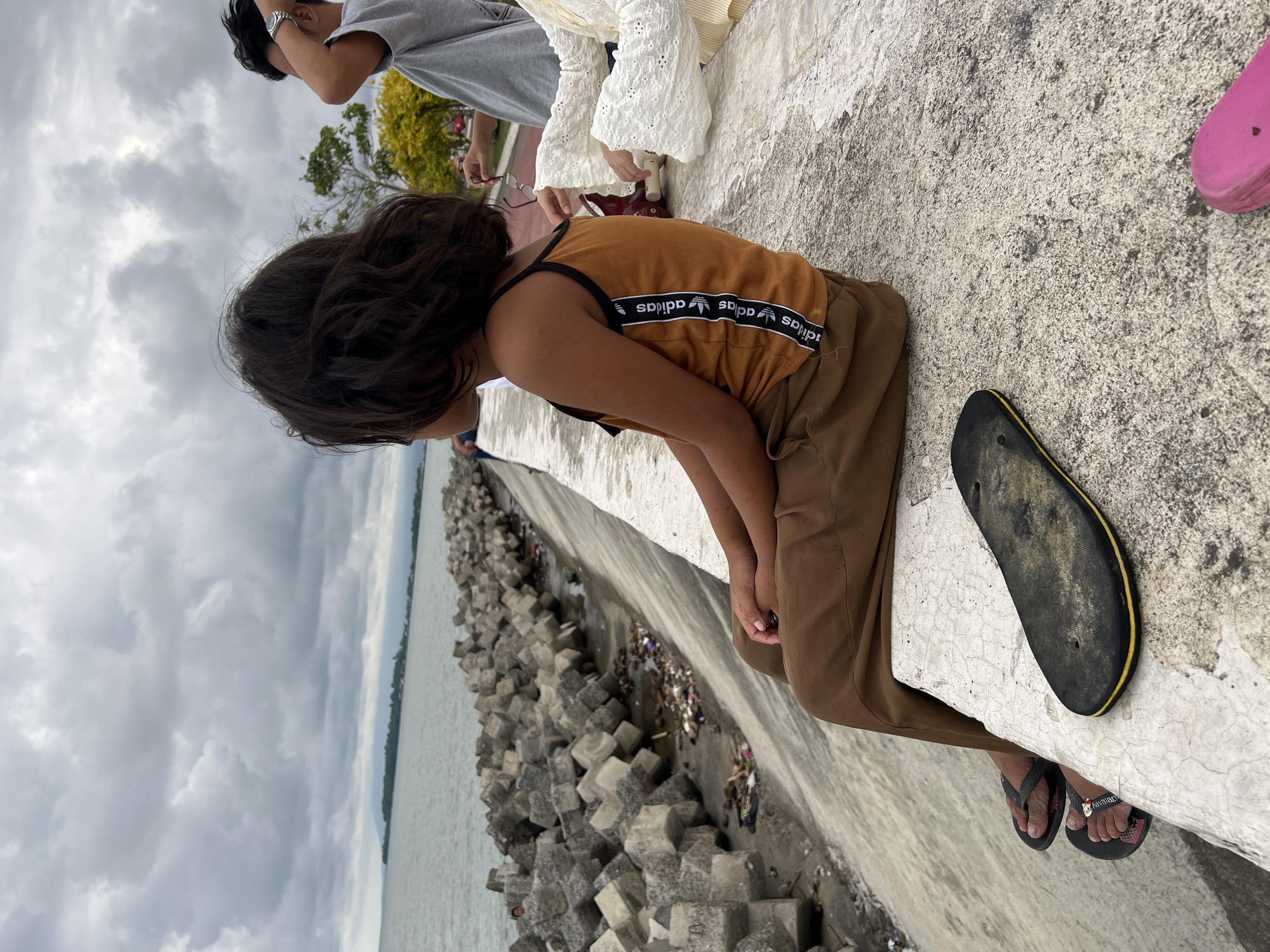 The image depicts a cloudy day scene at a coastal area in Davao City, Philippines, specifically along the seawall. In the foreground, a young child with dark, wavy hair is seated on the seawall, looking out at the water. They are wearing a tan-colored tank top with Adidas branding down the side and brown pants. A single black flip-flop rests on the seawall in front of them, and another pink flip-flop is partially visible to the right. To the right of the child, another person, partially visible, is wearing a white, lacy garment. In the mid-ground, concrete tetrapods form a barrier along the water's edge, with debris accumulating in the space between the tetrapods and the seawall. The sea stretches out to the horizon, meeting a cloudy sky. Further back, a person in a gray t-shirt is visible, with trees and a distant shoreline. The overall impression is one of a casual, public gathering by the sea under overcast weather conditions.
