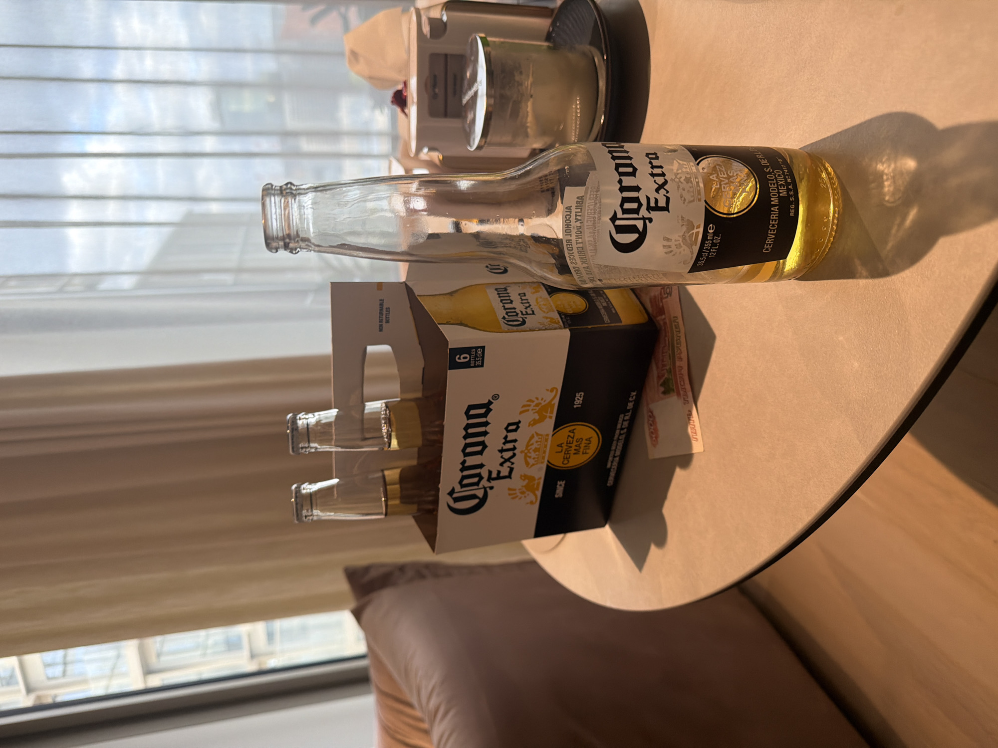 The image captures an indoor scene, likely a hotel room or apartment in Moscow, Russia, characterized by warm, golden sunlight streaming through a window. The main subjects are a half-full (or mostly empty) bottle of Corona Extra beer standing upright on a round, light-colored table, and a six-pack carrier of the same brand containing several other empty or nearly empty bottles.
The activity suggested is the consumption of beer, indicative of relaxation or leisure. The sunlight, casting long, distinct shadows from the bottle and other items on the table, points to either an early morning or late afternoon time of day, with the warm hue leaning towards late afternoon or early evening. In the background, visible through the window, are horizontal blinds, and a glimpse of an outdoor view can be seen beneath them. On the table further back, a metallic appliance, possibly a coffee maker or kettle, is visible. Part of a brown upholstered pillow or cushion is also visible in the foreground, suggesting a bed or sofa nearby.
Visible text on the main bottle includes "Corona Extra," "CERVECERIA MODELO, S. DE R.L. DE C.V. MEXICO," "REG. S.S.A. N°7417 "B"," "35.5 cl / 355 ml e," and "12 FL. OZ." The word "ALCOHOL" is visible in reverse. The six-pack carrier prominently displays "Corona Extra," "6 BOTTLES," "35.5 cle," "SINCE 1925," "LA CERVEZA MAS FINA," and "NON RETURNABLE BOTTLES." A small piece of paper near the six-pack shows the number "6000" and what appears to be "напитки," potentially indicating Russian text, which aligns with the provided location context.