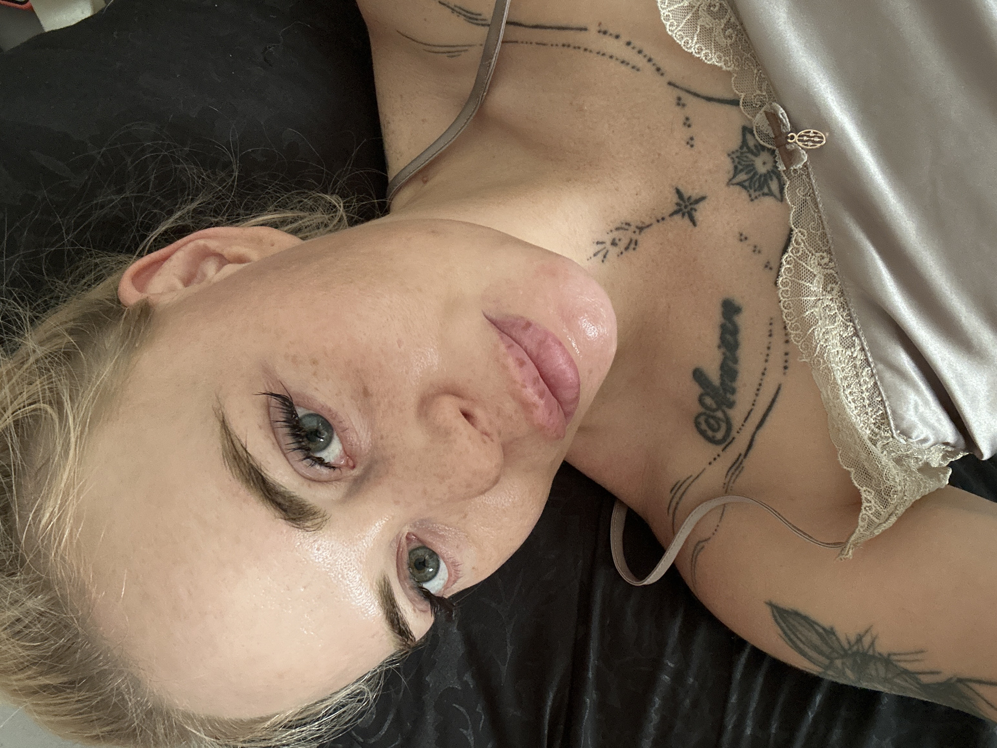 A close-up overhead shot shows a fair-skinned woman with blonde hair, lying down on a dark satin surface. She is looking up at the camera. Her eyes are green, and her eyelashes are long and dark. She has freckles across her nose and cheeks. Her lips are full and slightly parted. She is wearing a pale, silky camisole with lace trim. Several tattoos are visible on her upper chest and shoulder area. One tattoo on her chest appears to be script, and another on her shoulder depicts a stylized insect or moth.