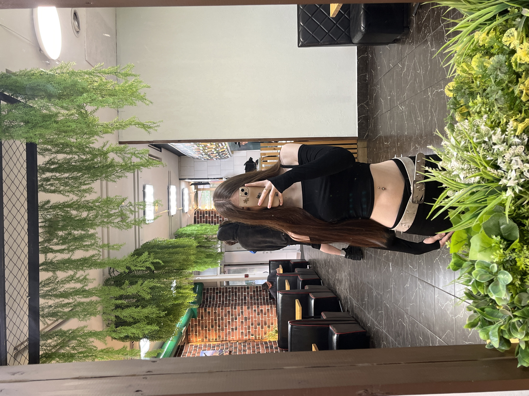 In a dimly lit establishment, possibly a fast-food restaurant or cafe, a young woman with long brown hair is taking a mirror selfie. She is wearing a black off-the-shoulder crop top with long sleeves, black pants, and a beige belt. Her belly button piercing is visible. In the reflection, we see rows of black booth seating with red trim, brick walls, and hanging artificial greenery. Two other individuals are visible in the background, one wearing a dark hoodie and the other a dark t-shirt. The floor is tiled in a dark marble pattern. The presence of Russian signage, like the word "КАФЕ" (cafe) visible on a menu board in the reflection, suggests the location is in Saint Petersburg, Russia. Artificial plants are also visible in the foreground, obscuring the bottom part of the mirror.