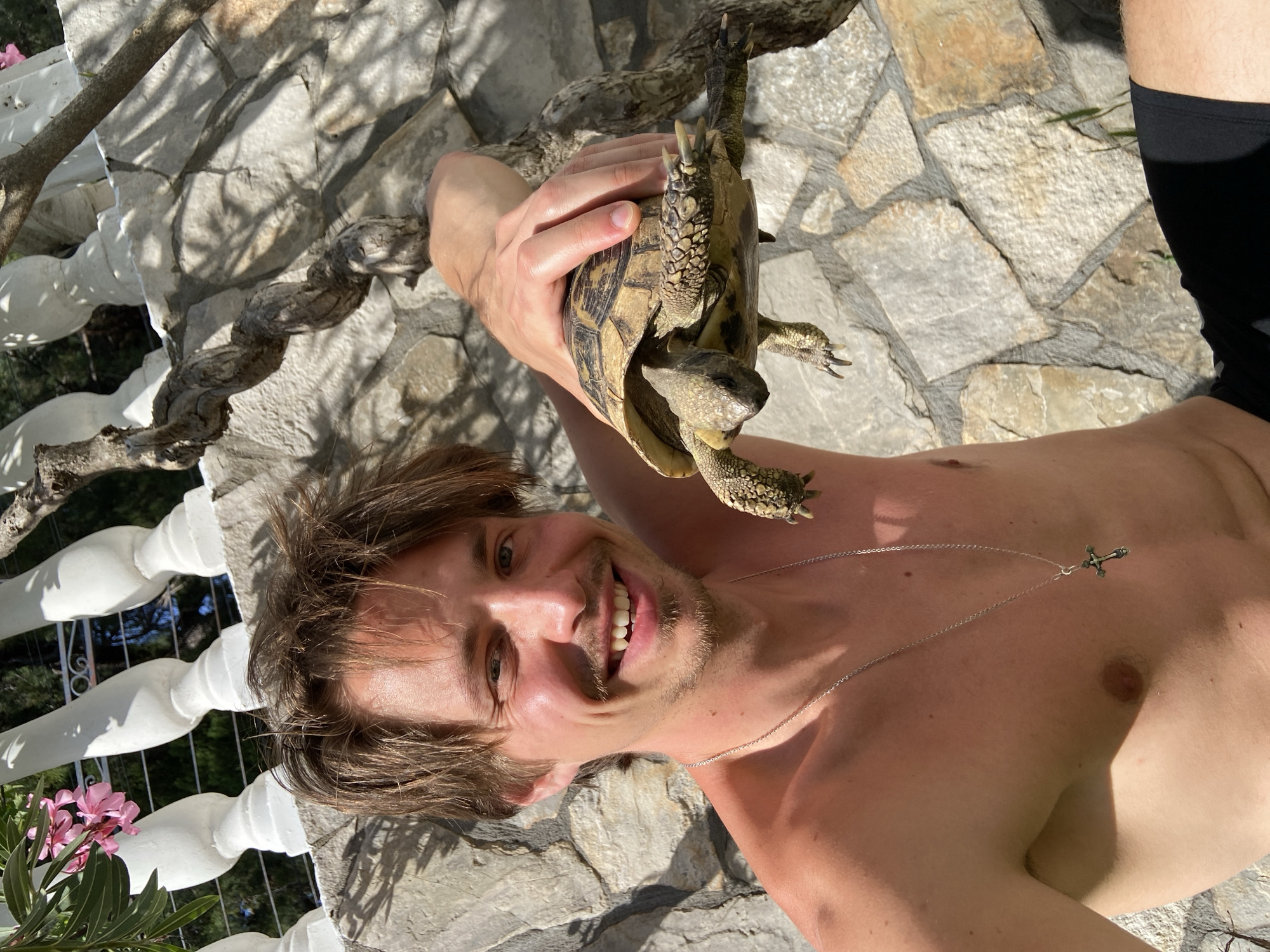A man with brown hair and a slight mustache, wearing a silver necklace with a cross pendant, is holding a tortoise in his hand, near his head. The man is shirtless and wearing dark shorts or swimwear. He is smiling and looking at the camera. The background includes a stone wall or patio and a tree branch with a white railing and flowers to the side. The location is Šušanj, Montenegro.