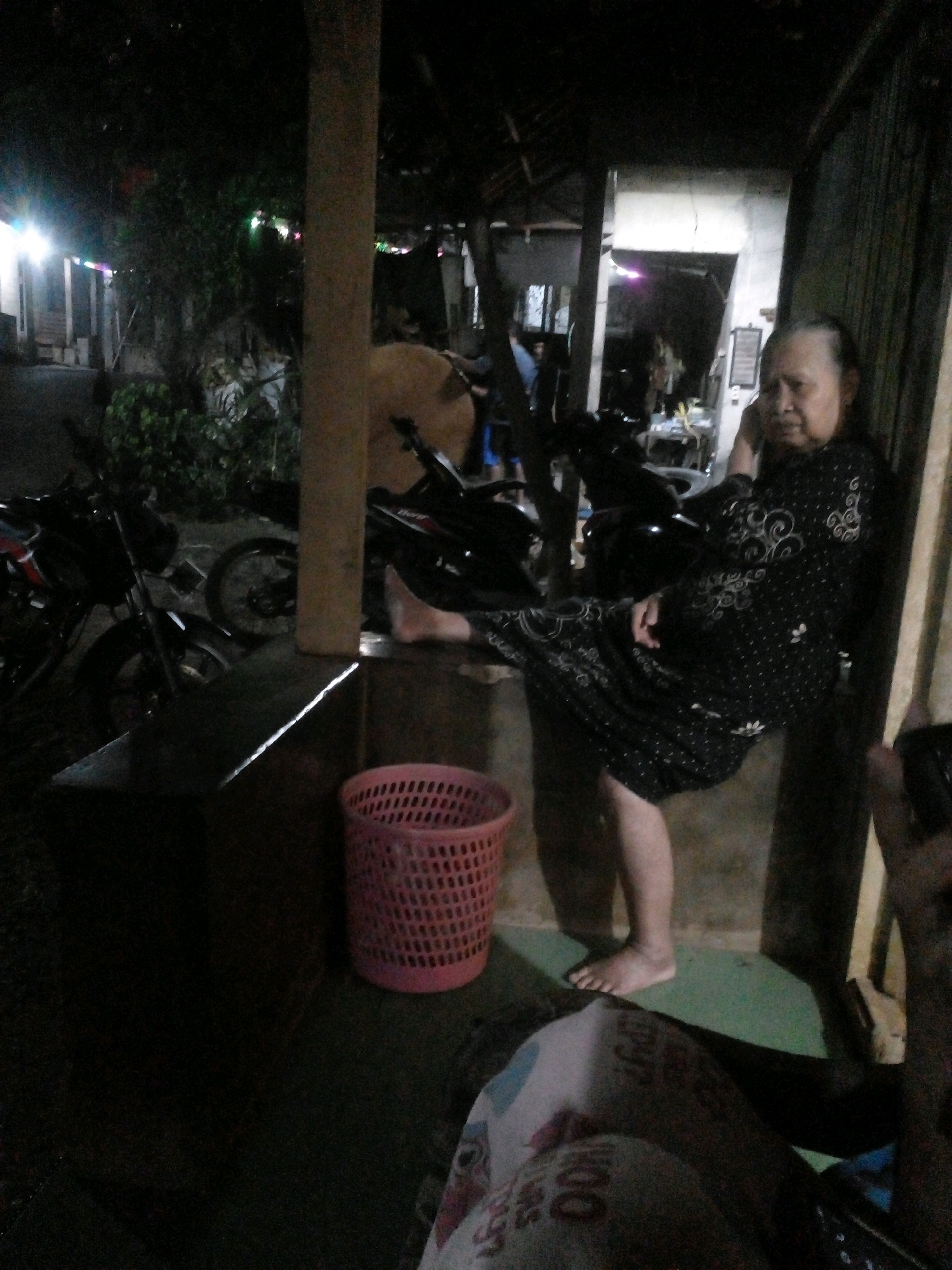 In Pungangan, Indonesia, a woman is seated, resting her right hand on her face while holding a mobile phone to her ear, suggesting she is engaged in a conversation. She is wearing a black dress adorned with white floral patterns. Her left leg is bent at the knee and extended outwards, with her left foot resting on the floor. To her left, a pink plastic basket is visible on the ground. Behind the woman, several motorcycles are parked, indicating an outdoor or semi-outdoor setting, possibly a residential yard or a communal space. The lighting is dim, suggesting it is nighttime, with artificial lights illuminating parts of the scene. The environment appears to be a modest, possibly rural or suburban area, with a building structure visible in the background.