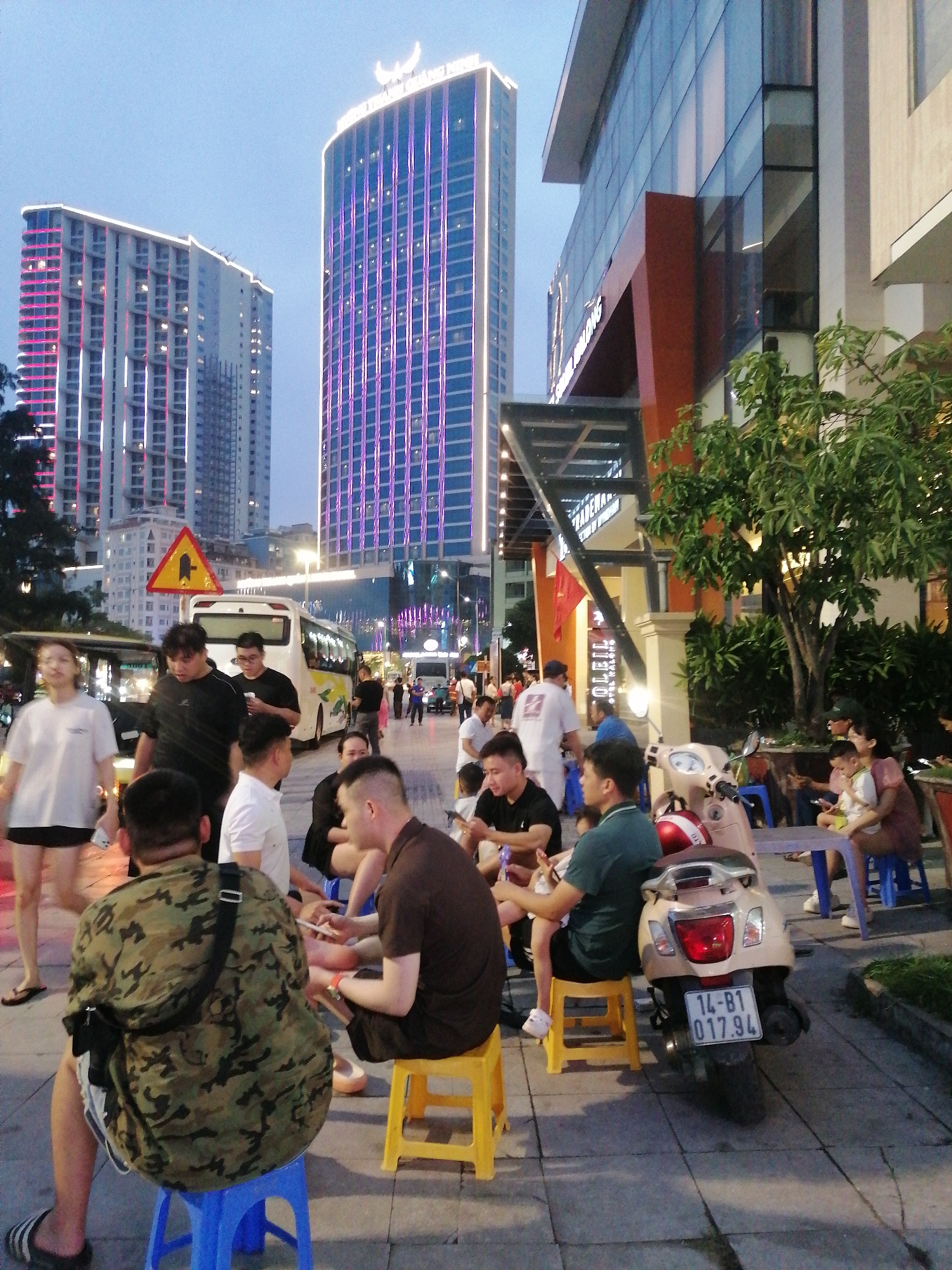 The photograph captures an outdoor scene in Bai Chay Ward, Vietnam. Several individuals are seated on small, colorful plastic stools along a sidewalk, engaged in conversation or activities. A beige scooter with license plate "14-B1 017.94" is parked nearby. Pedestrians are walking in the background. The backdrop features modern high-rise buildings with decorative lighting, including one with a crescent-shaped light on top. A traffic sign is also visible, along with other vehicles. The environment indicates an urban setting with businesses and public spaces.