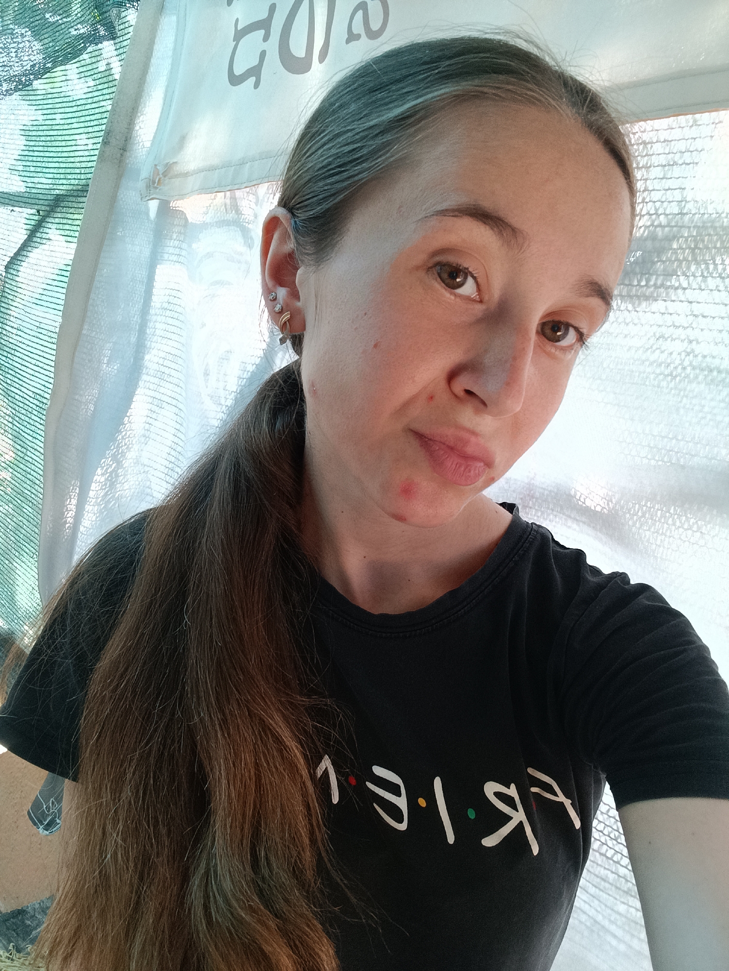 A young woman with long brown hair tied in a ponytail is pictured in an outdoor setting, possibly in Vypasne, Ukraine. She is wearing a black t-shirt with the word "FRIENDS" emblazoned on it in white lettering. The background consists of a white fabric structure, likely a canopy or tent, with some green netting visible on the left. The woman has light skin and brown eyes, and a few red blemishes are visible on her face. She is looking towards the camera with a neutral expression. The lighting suggests it is daytime, with natural light illuminating the scene.