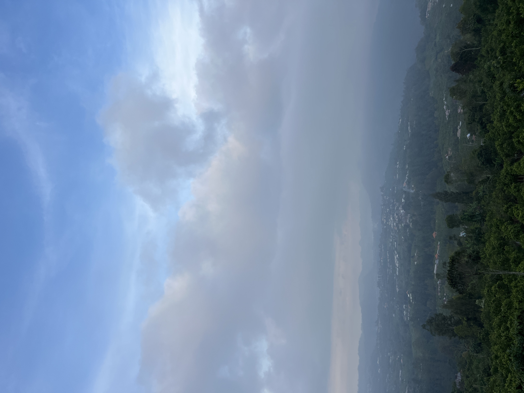 The image shows an elevated view of a landscape in Kintamani, Indonesia. The sky, partially obscured by clouds, transitions from blue to grey. Below the clouds, a hazy vista reveals a settlement nestled in a valley. Scattered buildings and vegetation are visible across the landscape. The foreground is marked by dense greenery, possibly forest. No people are visible in the image.