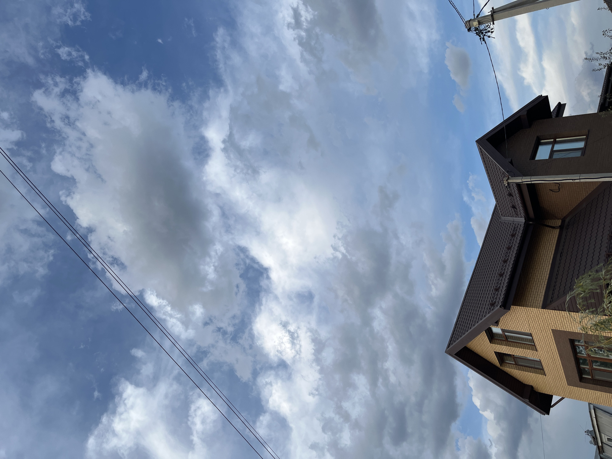 The image displays a two-story house with a dark brown roof and light brown brick exterior, situated in the city of Shumeyka, Russia. The house has several windows with dark frames. Above the house, a blue sky is visible, populated with scattered white and grey clouds. Power lines run diagonally across the upper portion of the frame, and a utility pole with wires is present on the right side of the image. Some tree branches are also visible in the lower right corner.