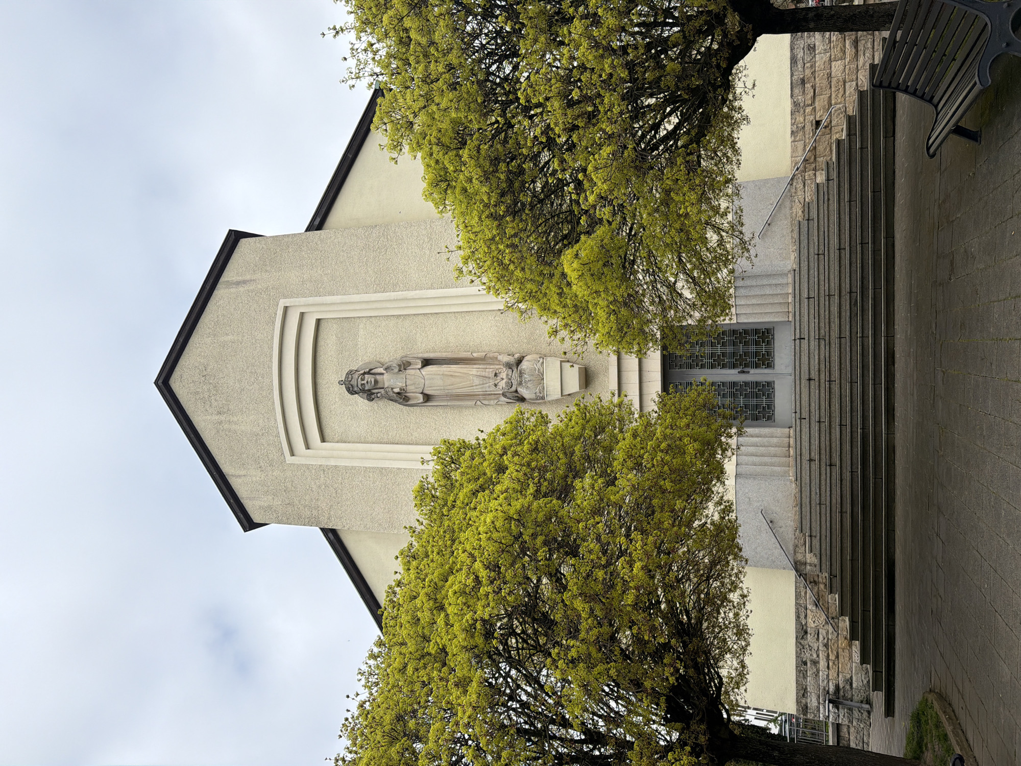 The image depicts the façade of the church of Saint-Henri, located in the city of Esch-sur-Alzette, Luxembourg. The building is characterized by a gabled structure, with a neutral beige exterior. Above the entrance is a stone relief depicting a figure lying down in repose. Two trees with vibrant green foliage partially obscure the view of the church. A staircase ascends toward the entrance, flanked by a paved area and a bench on the right side. The sky is overcast.