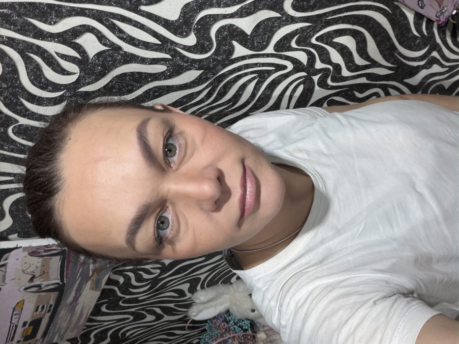 A woman poses for a selfie, reclining against a black and white zebra-patterned surface. She wears a white t-shirt. Her face is centered, with a neutral expression, light skin, and green eyes. Light brown hair is pulled back. A glimpse of a necklace is visible at her collarbone. In the bottom left, a drawing or a pattern is visible. A small white stuffed animal with purple flowers lies to her right.