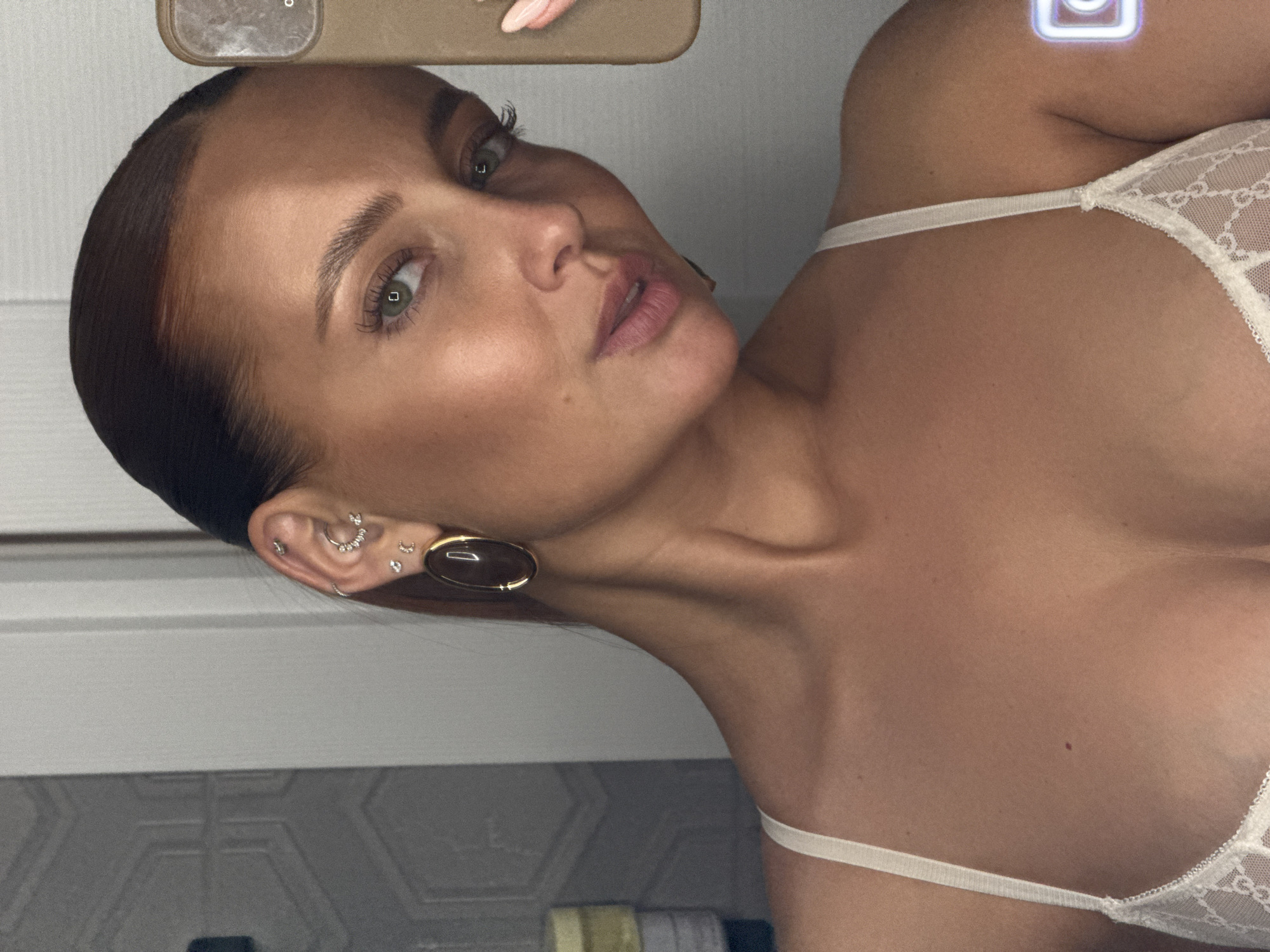 A woman with fair skin, green eyes, and dark hair pulled back is positioned in the center of the frame, angled slightly to the side. She is wearing a light-colored lingerie top with white straps, and several earrings in her left ear. The background appears to be a simple, tiled bathroom setting. There is no indication of the location being Chaikovsky, Russia.