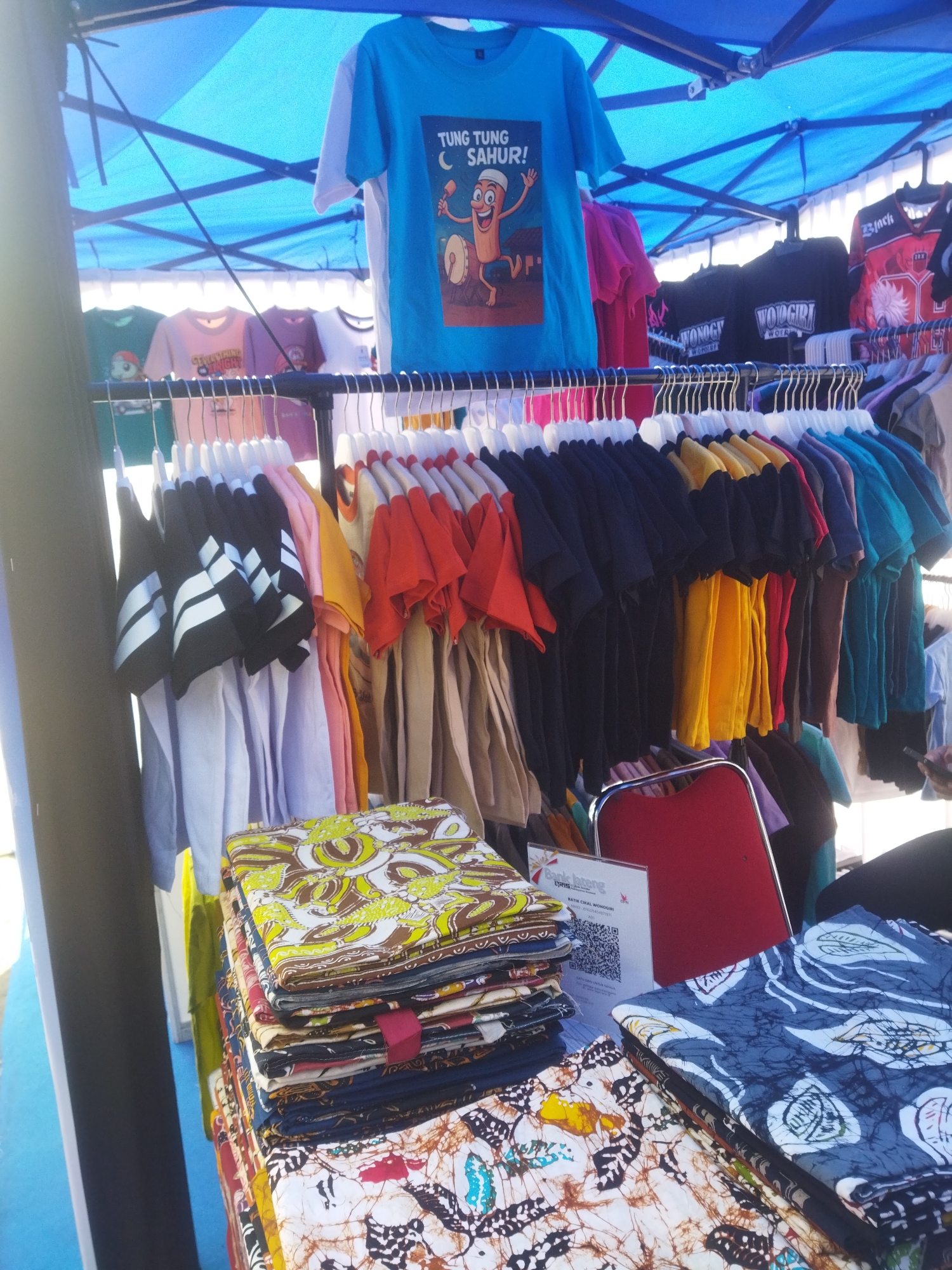 The image depicts a street stall in Giripurwo, Indonesia, displaying a variety of clothing items. A blue canopy provides shade over the stall. Several t-shirts hang on a rack, with one prominent blue t-shirt featuring a cartoon character and the text "TUNG TUNG SAHUR!". Below the hanging t-shirts, stacks of folded fabric, likely batik, are arranged on a table. One stack displays a pattern of green, yellow, and brown motifs, while another shows a blue and white leaf pattern. A red chair is visible behind the fabrics. The overall scene suggests a market or a retail setup outdoors.