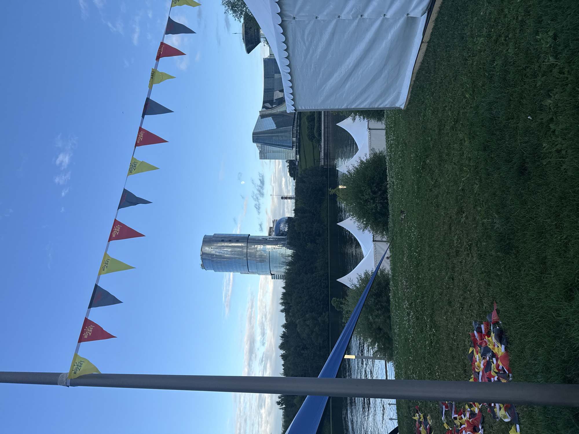 The image depicts an outdoor scene with a clear blue sky, partially obscured by a few wispy clouds. A string of triangular flags, alternating in colors of red, yellow, and dark gray, hangs across the frame from the left. Some of these flags have text that appears to read "RUS • SVET". White tents are visible in the upper right and mid-ground, suggesting an outdoor event or gathering.
In the background, a modern glass-covered skyscraper and other buildings rise above a line of trees. A body of water, likely a river or lake, is present in the mid-ground, reflecting the sky and surrounding environment. The foreground consists of a grassy area, where a collection of smaller flags, also in red, yellow, and dark gray, are clustered on the ground. A blue tarp or fabric is stretched diagonally across the lower right portion of the image. The overall lighting suggests it is either early morning or late afternoon. The setting appears to be a park or waterfront area in Khimki, Russia. No people are clearly visible in the frame.