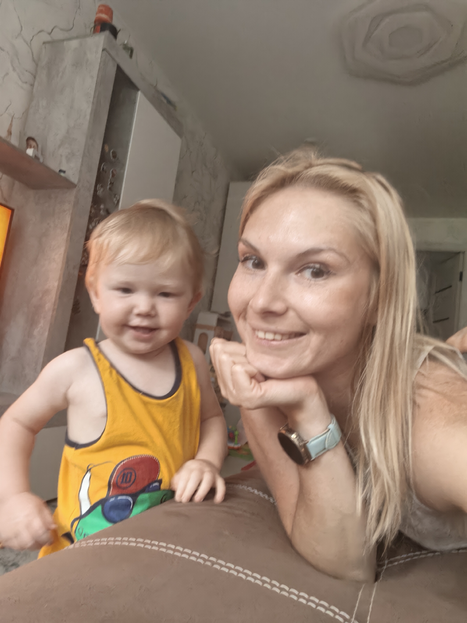 The image shows a young Caucasian child and an adult Caucasian woman indoors. The child, appearing to be a toddler, is wearing a yellow tank top with a graphic print and is positioned on the left side of the frame. The woman is on the right side, resting her chin on her hand and looking towards the camera with a smile. She has blonde hair and is wearing a light-colored top and a watch on her left wrist. They are both in a room with a brown upholstered surface in the foreground. In the background, there is a wall with textured wallpaper and a light-colored cabinet. The scene suggests a casual, domestic setting. No specific location within the city of Sterlitamak, Russia, can be identified from the image alone.