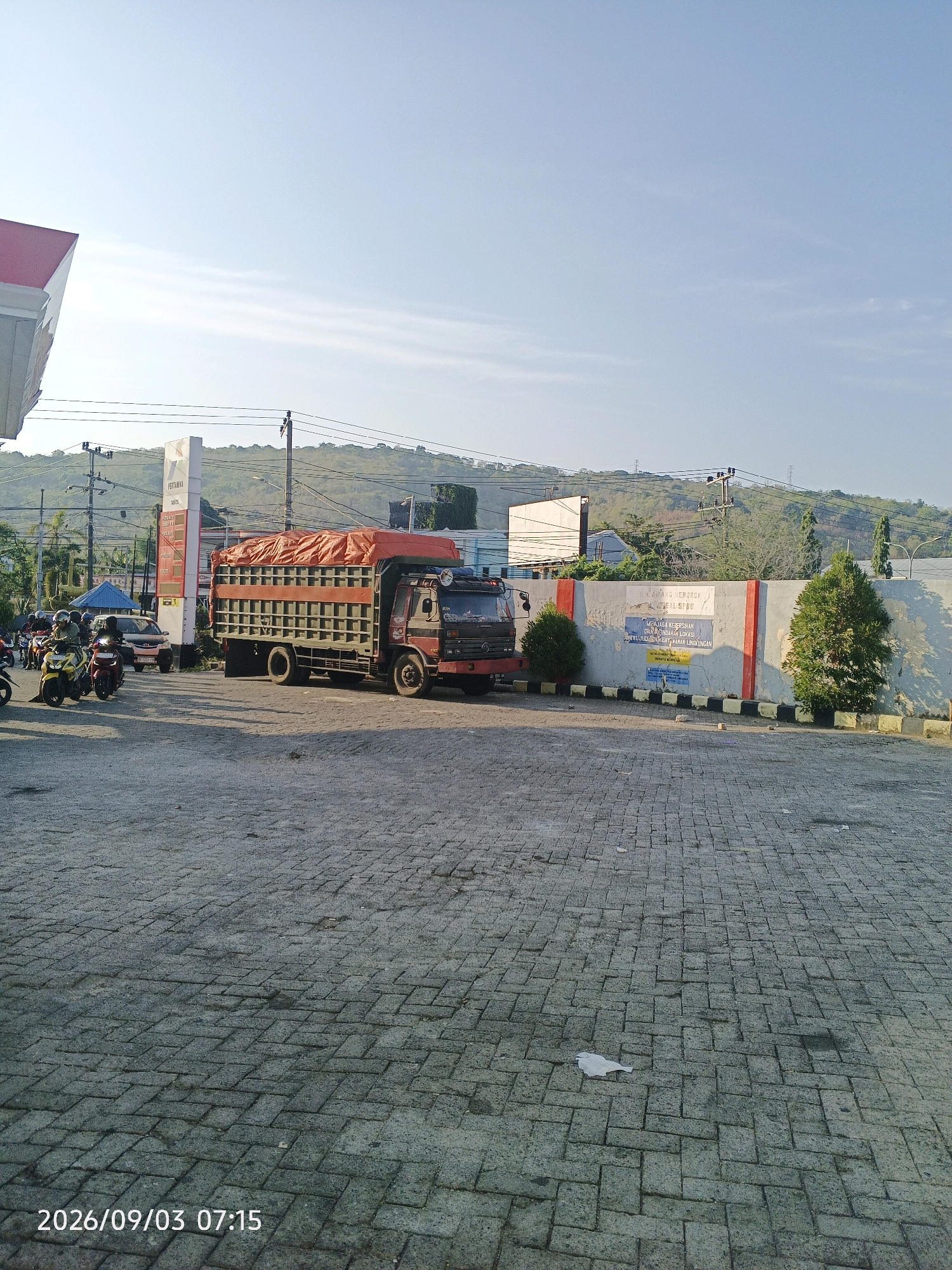 The image depicts an outdoor scene at a Pertamina gas station in Pare-Pare, Indonesia, as indicated by the visible Pertamina sign and the text "2026/09/03 07:15" which suggests the date and time. The foreground is paved with interlocking grey bricks. A red-canopied structure, likely part of the gas station's canopy, is visible on the far left.
In the mid-ground, a large truck with a red tarp covering its cargo is parked on the pavement. To the left of the truck, several motorcycles are seen, with riders and passengers. One yellow motorcycle with two riders is prominent. Behind the motorcycles, a dark-colored car is visible. A tall white pillar with red accents, also part of the Pertamina signage, stands near the truck.
The background features a concrete wall with red and white striped sections and some signage. Beyond the wall, a lush green hillside rises under a clear, light blue sky with faint wispy clouds. Power lines and poles are also present in the background, suggesting an urban or semi-urban environment. Two small evergreen trees are planted in front of the wall. The lighting indicates it is morning, with the sun casting shadows on the ground. The overall scene is of a busy, functional gas station.