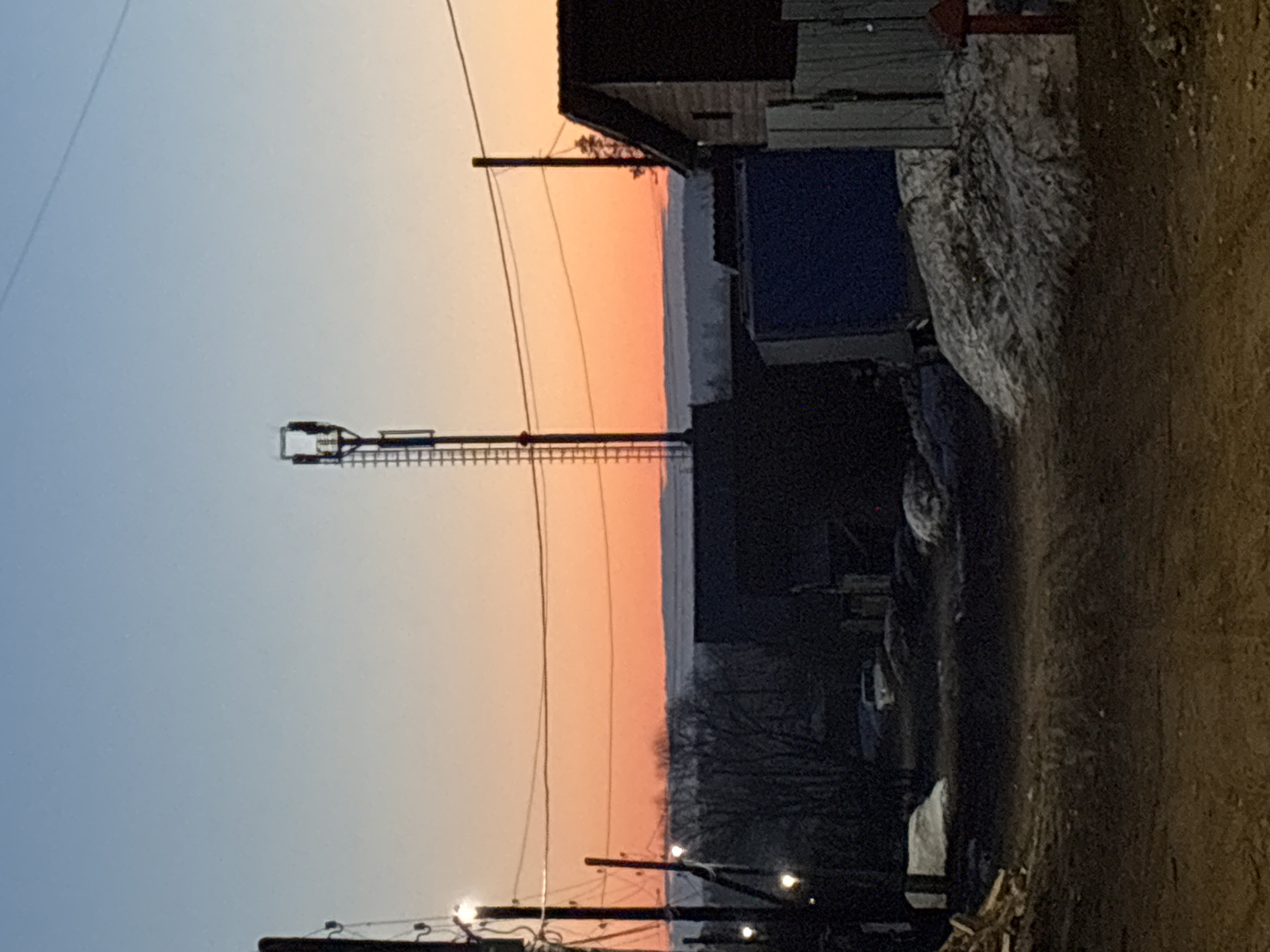 The image shows a street in Chaikovsky, Russia at sunset. The sky is a gradient of blue to orange. A transmission tower stands tall against the horizon, while power lines crisscross the lower portion of the image. To the right, there is a building and snow on the ground. The road appears unpaved and runs along the right edge of the frame.