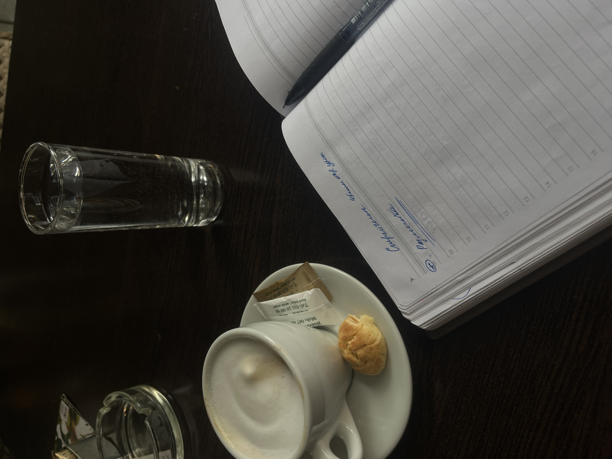 The scene depicts a tabletop in a cafe located in Budva, Montenegro, as indicated by text on a sugar packet displaying "Budva, MNE" and a local phone number. A dark wooden table surface serves as the setting. On the left, a white ceramic cup containing a cappuccino with foamed milk sits on a matching saucer. Beside the cup, a small, golden-brown pastry with a broken piece is present. Two sugar packets, one open brown sugar packet and one closed white sugar packet, lie on the saucer. To the right, an open notebook with lined pages and numerical markings (09-16) on the margin displays handwritten Cyrillic text in blue ink, including "Сиријусом 45мин. отв. урок" and "Родитељи". A black ballpoint pen, marked "72% RECYCLED PLASTIC," rests across the open notebook pages. A tall, clear glass filled with water stands in the upper center of the frame. In the upper left corner, a clear glass ashtray, mostly empty save for a small piece of paper, is visible. No individuals are present in the image.